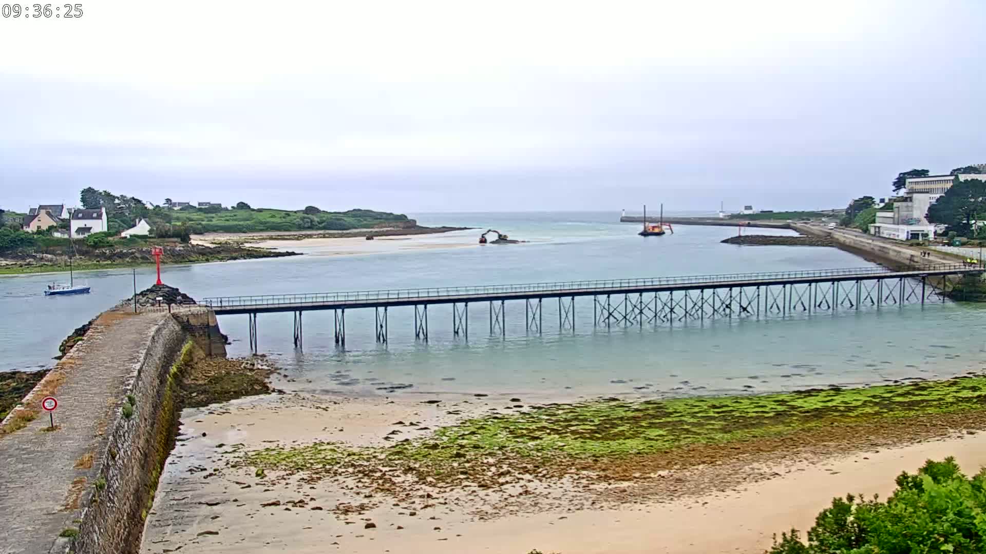 A long pier stretches across shallow, calm water toward a small, sandy beach and a distant shoreline on an overcast day.