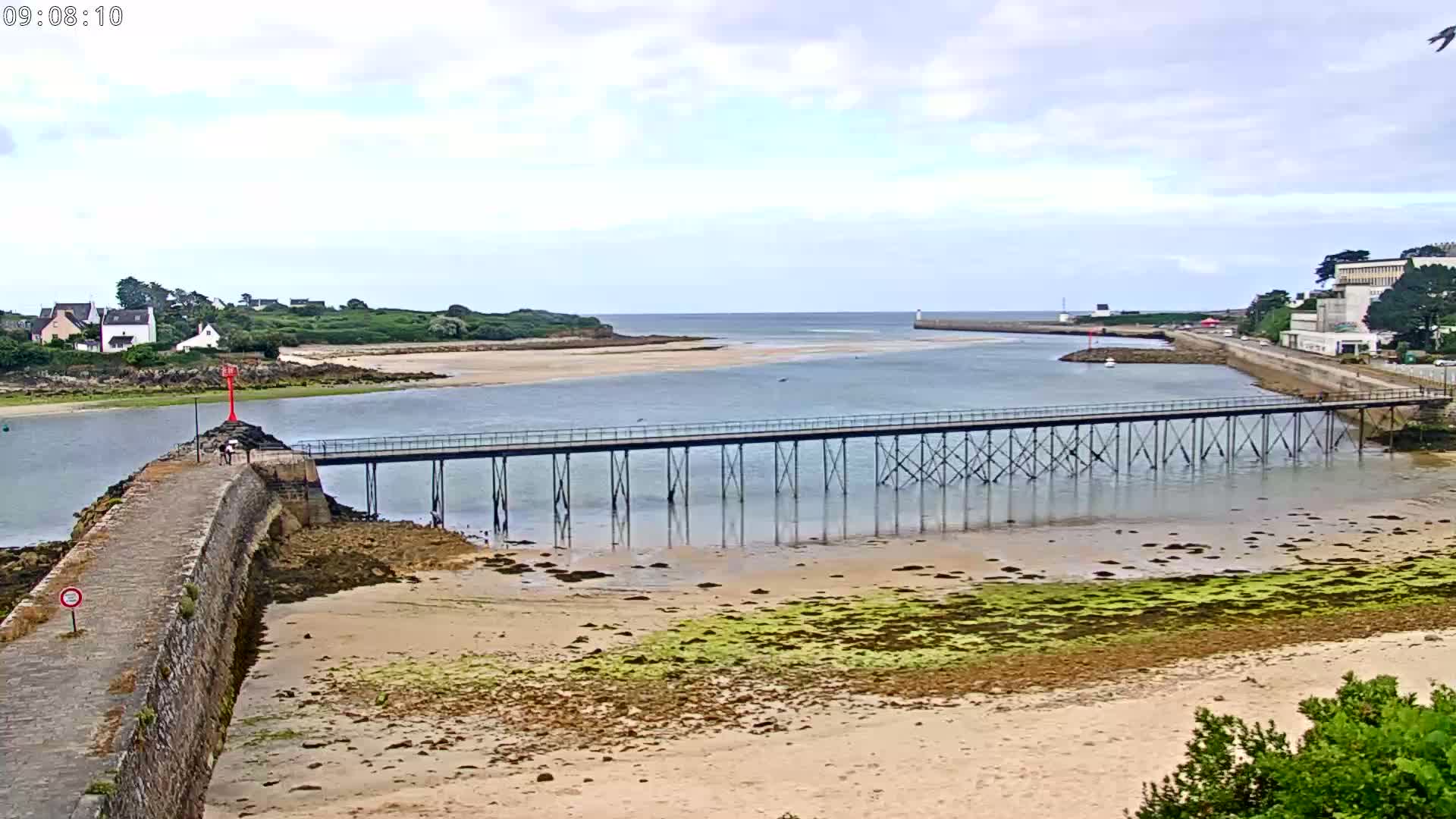 A long pier extends over shallow, calm water, connecting a stone jetty to a sandy shore lined with green algae, with a small village and buildings visible in the background under a partly cloudy sky.