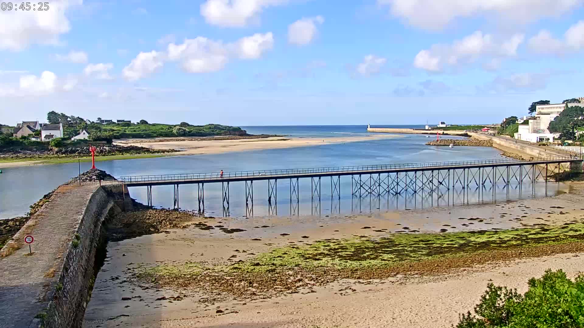 A long pier stretches across a calm body of water on a partly cloudy day, connecting a sandy beach to a small town with buildings and green hills.