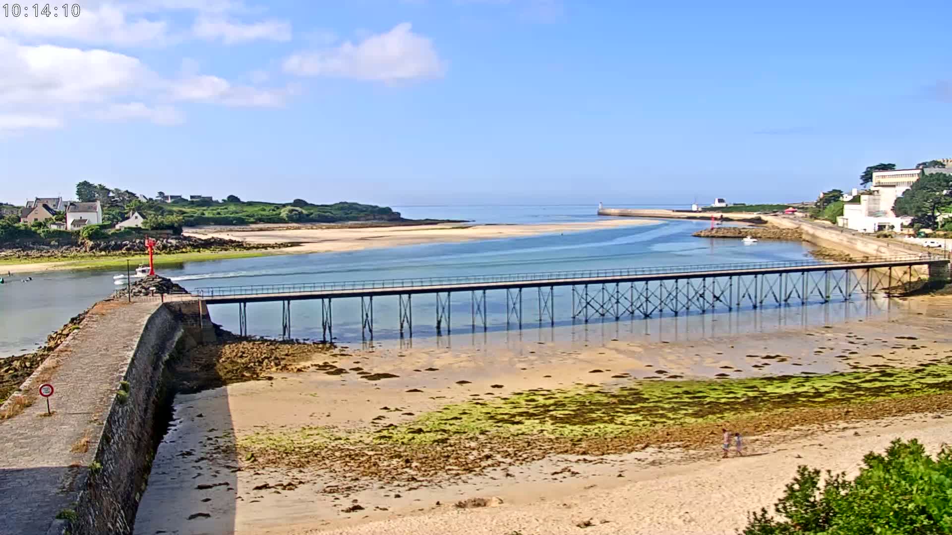 A long pier stretches across a shallow, sandy inlet bordered by a stone wall and low-lying buildings, under a mostly sunny sky.