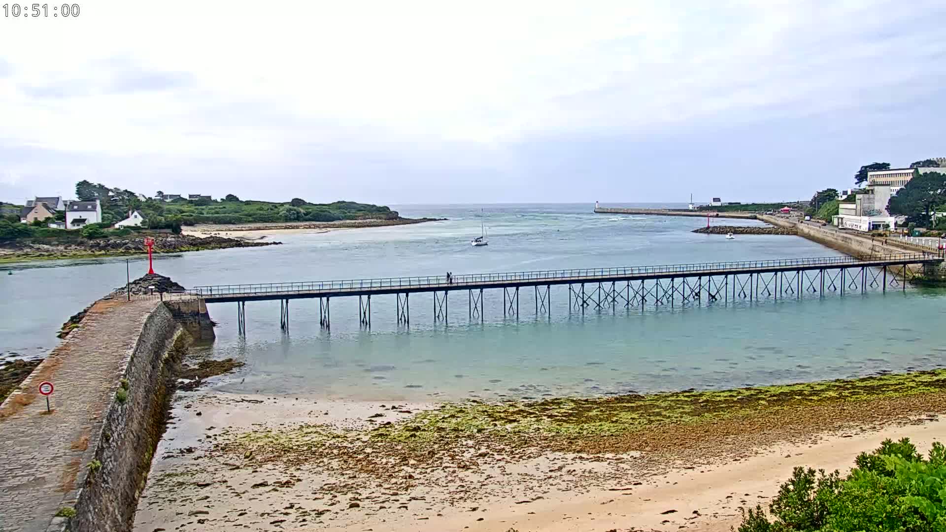 A long pier extends over calm, light-green water toward a small island under an overcast sky, with a sandy beach visible in the foreground.