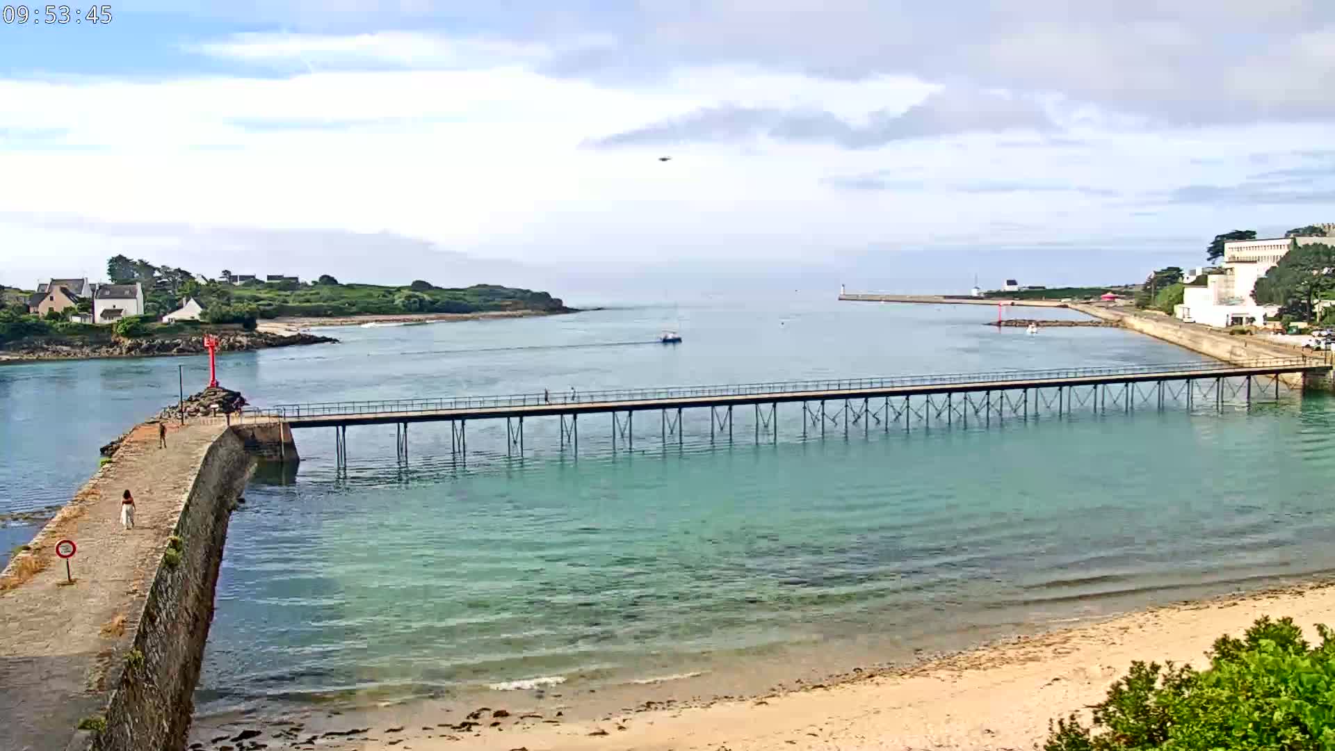 A long pier extends over calm, clear turquoise water toward a small island and distant buildings under a partly cloudy sky.