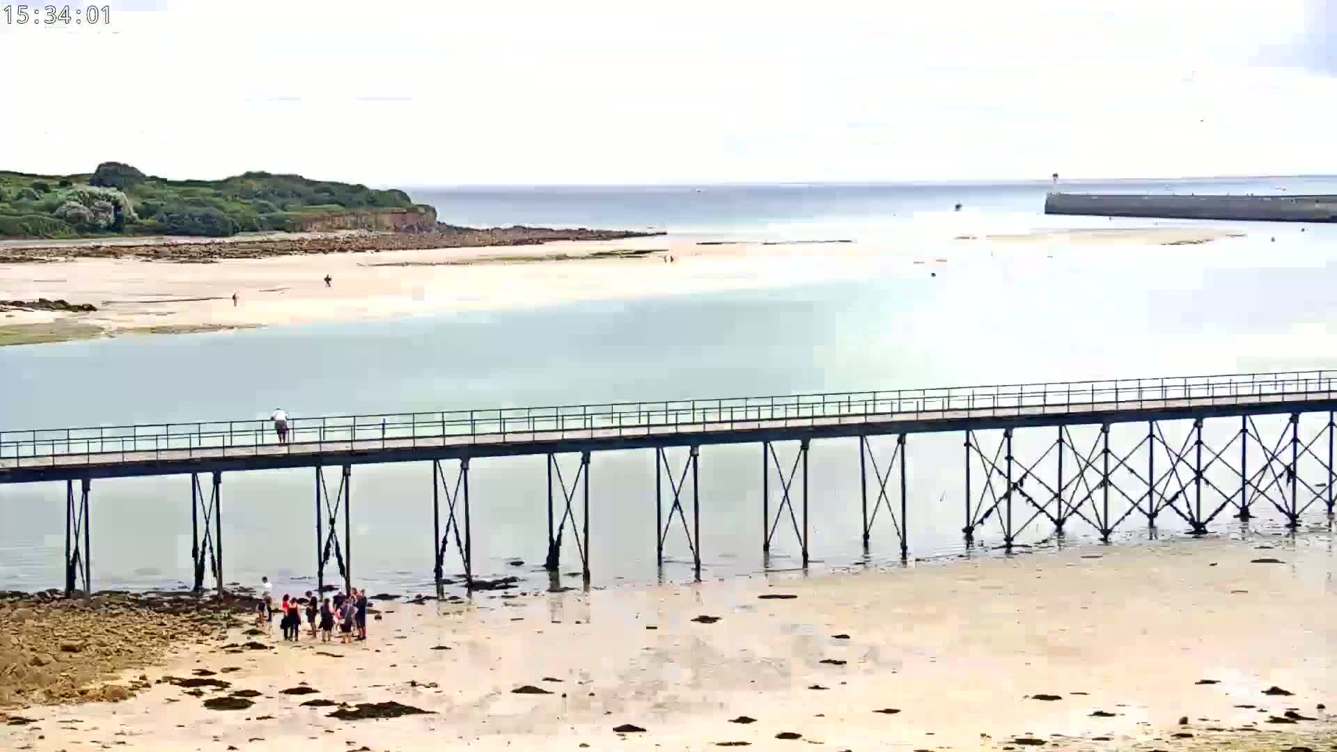 A long pier extends over calm, clear turquoise water toward a small island and distant buildings under a partly cloudy sky.