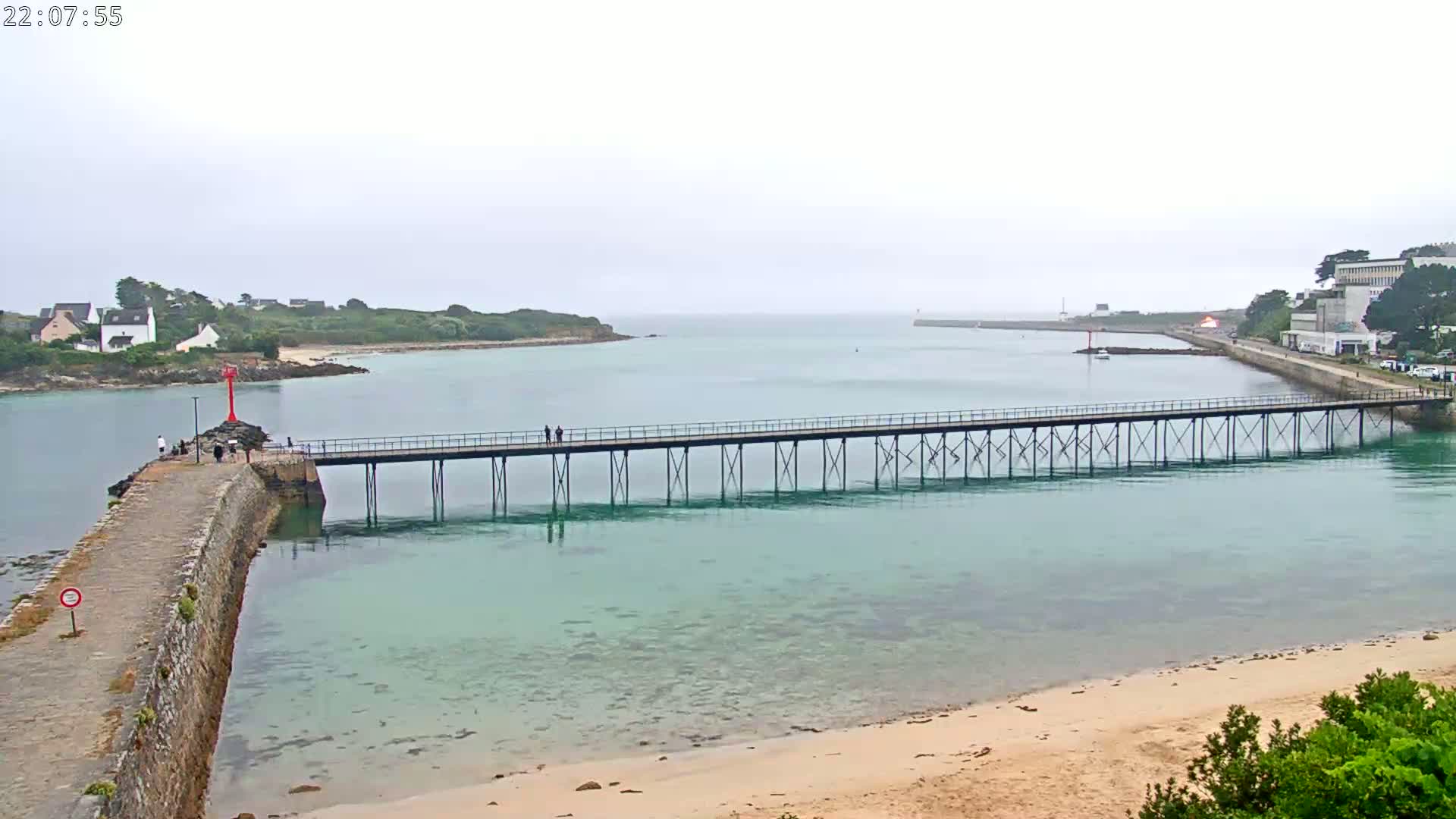 A long pier extends over calm, clear turquoise water toward a small island and distant buildings under a partly cloudy sky.