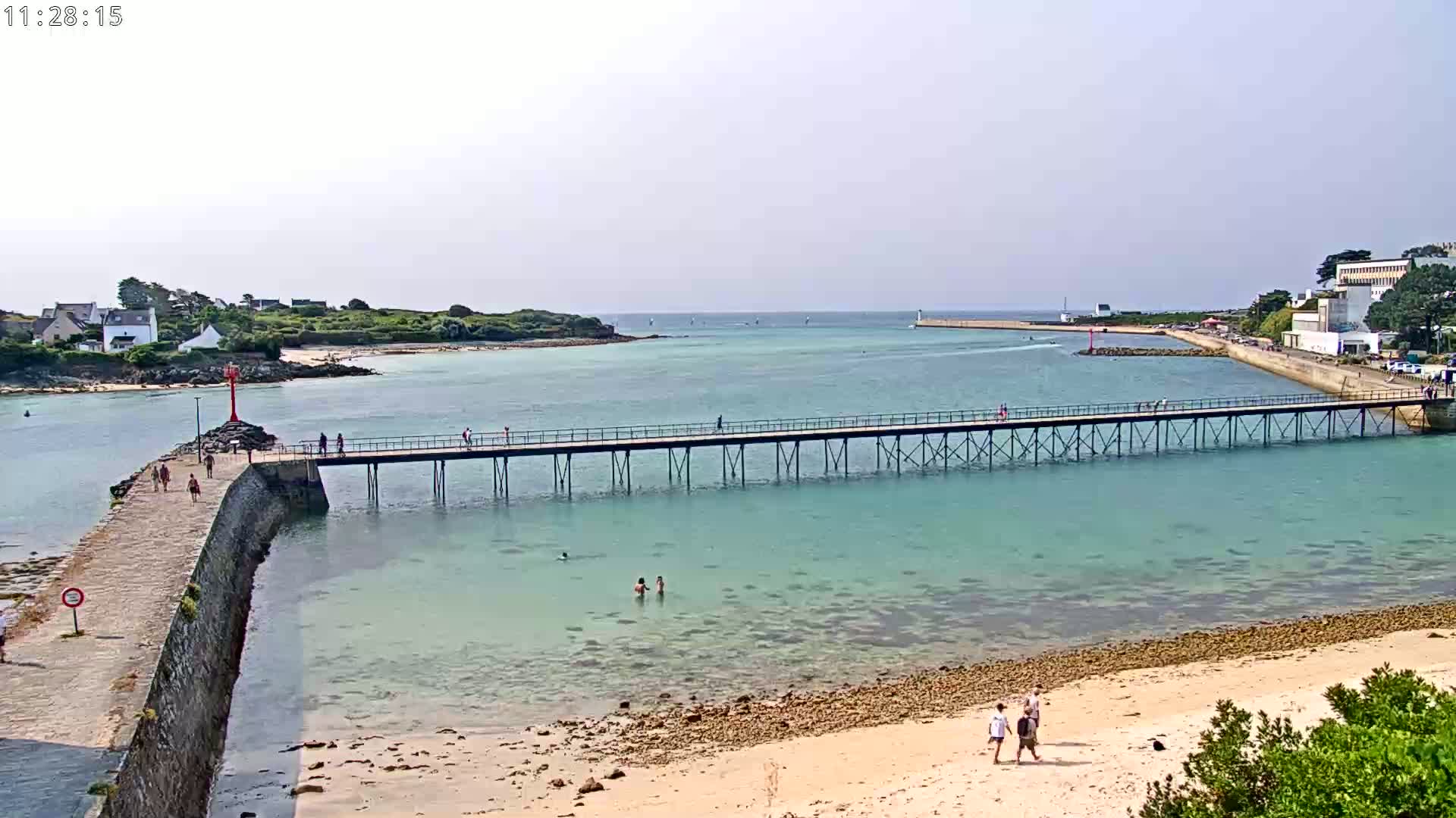 A long pier extends over calm, clear turquoise water toward a small island and distant buildings under a partly cloudy sky.