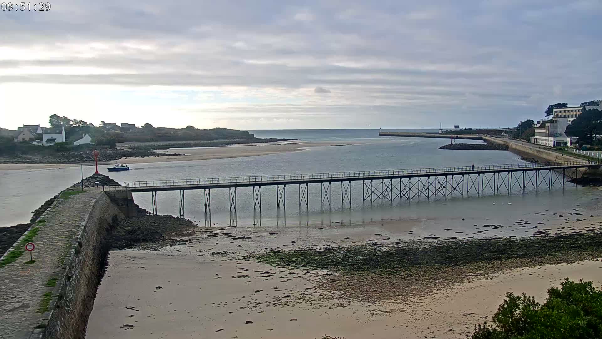 Under a cloudy sky, a long pedestrian pier stretches across a wide, sandy tidal inlet revealing exposed seabed, flanked by a stone breakwater with a red beacon, and coastal buildings on both sides, with a small boat visible in the water.