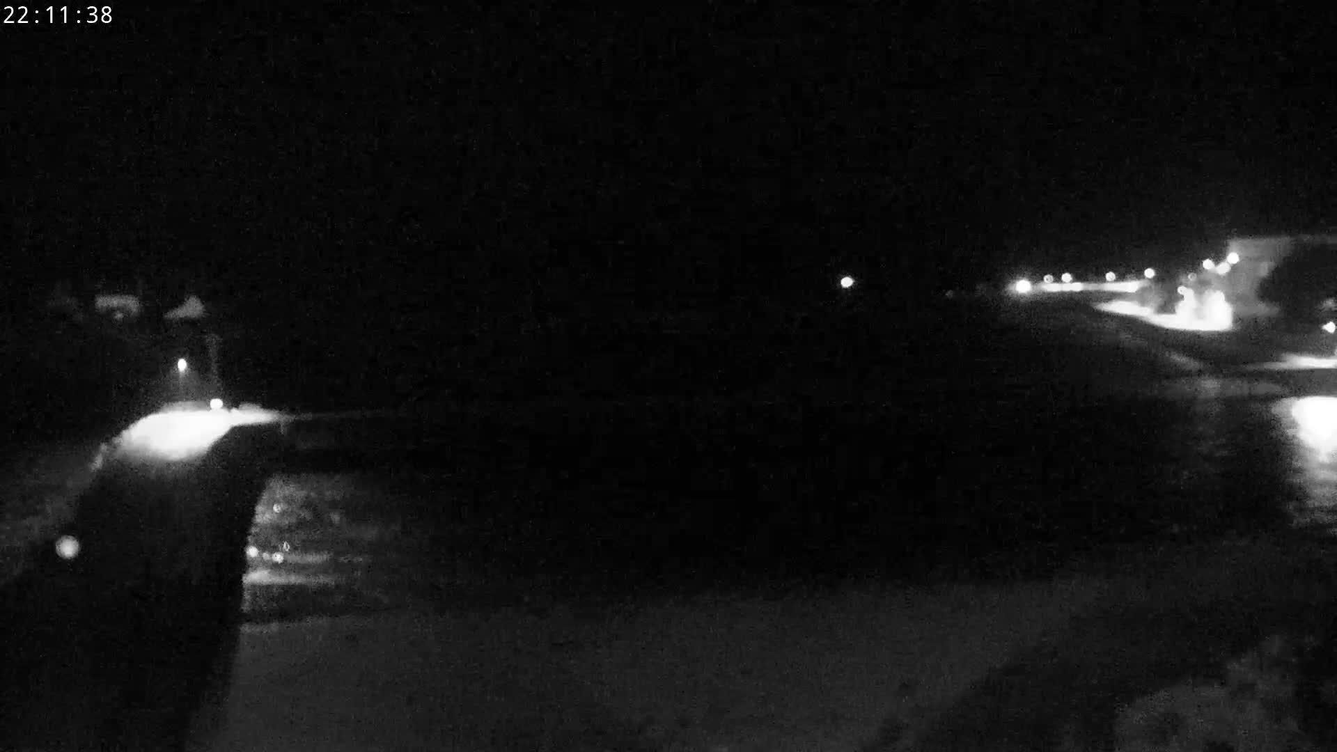 The image captures an overcast coastal scene with a long pier spanning a wide tidal river mouth or inlet, bordered by a stone causeway, sandy shores, distant buildings, and is viewed through a camera lens obscured by rain or moisture.