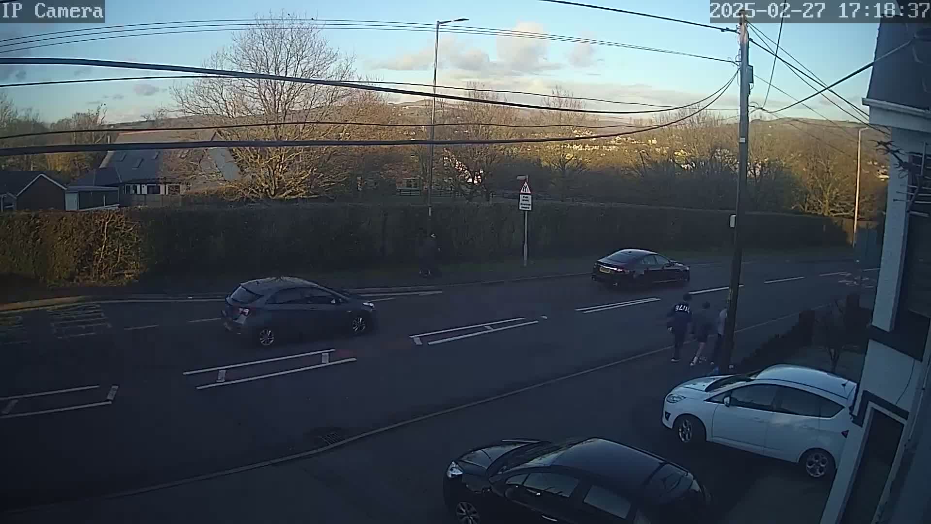 A street scene at dusk shows several cars parked and driving on a road, with pedestrians and a hedge in the background under a mostly clear sky.