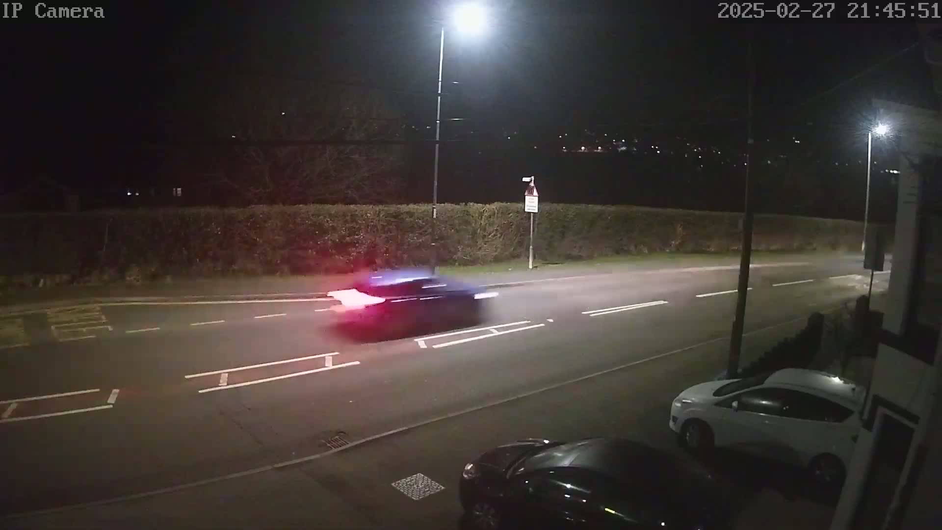 A dark-colored car drives down a road at night, past a parked white car, under bright streetlights.