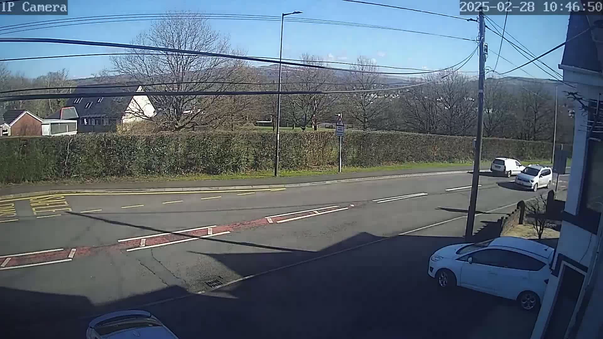 A sunny day shows a residential street scene with three white cars, one parked and two driving, and a hedgerow separating the road from houses and trees in the background.