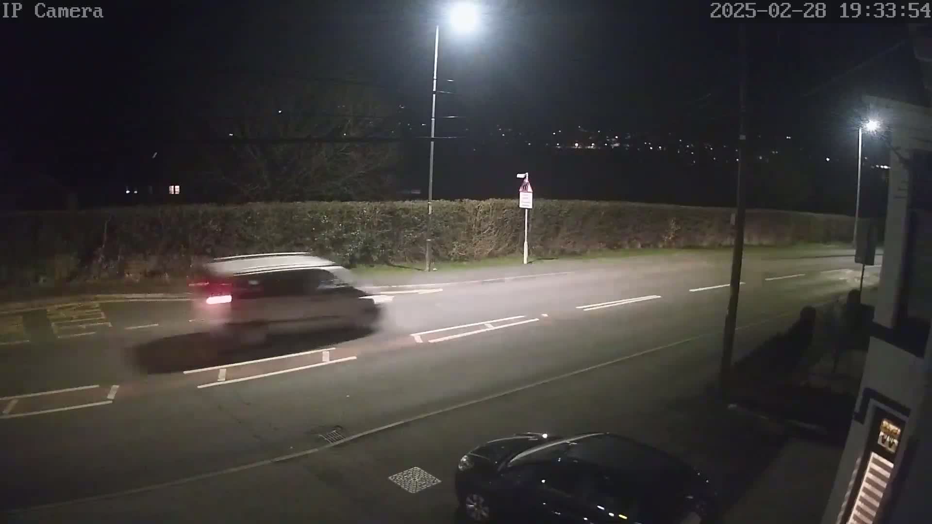A light-colored SUV drives away at night on a dark road,  underneath streetlights.
