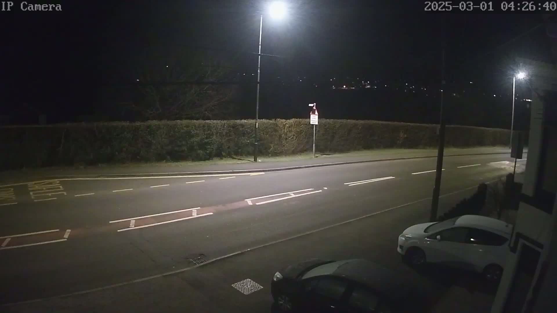 A nighttime street scene shows a road with cars parked along the side, lit by streetlights, with a hedge and distant lights visible in the background under a dark sky.