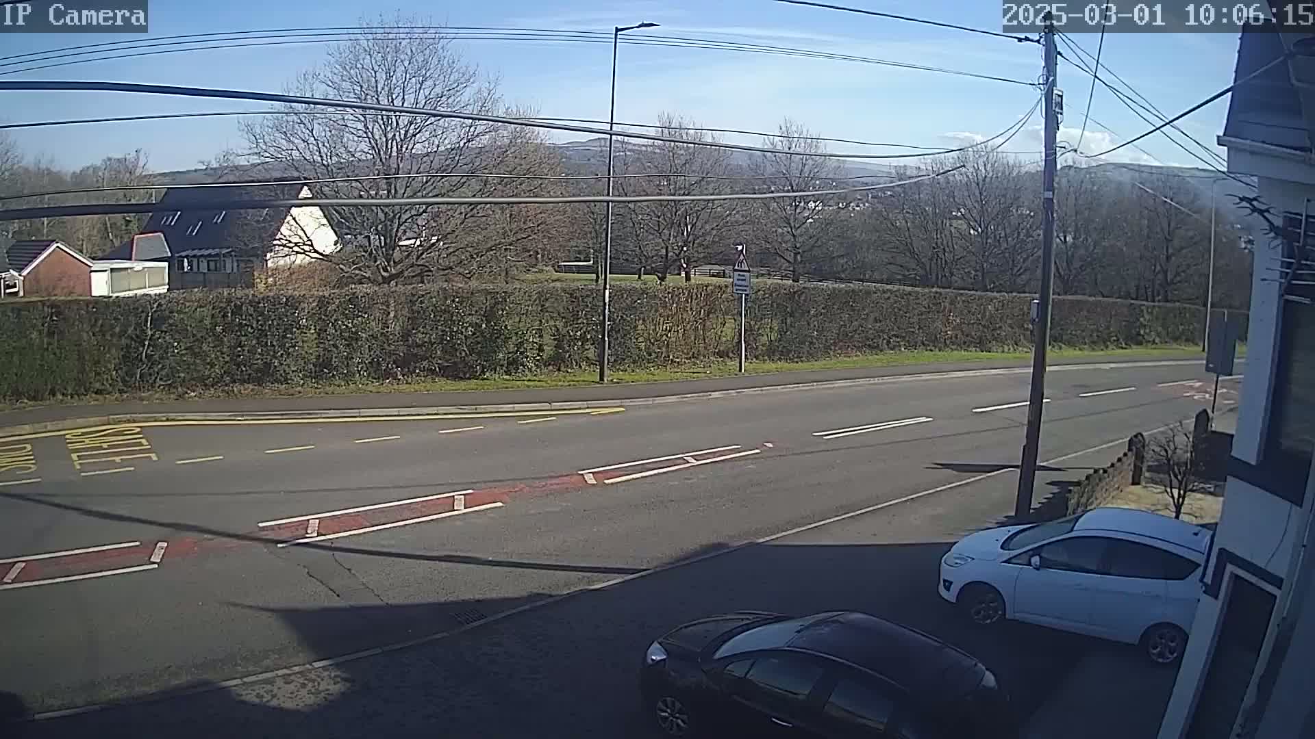 A sunny day reveals a road intersection with two cars parked nearby, bordered by a hedge and trees, with distant hills visible in the background.