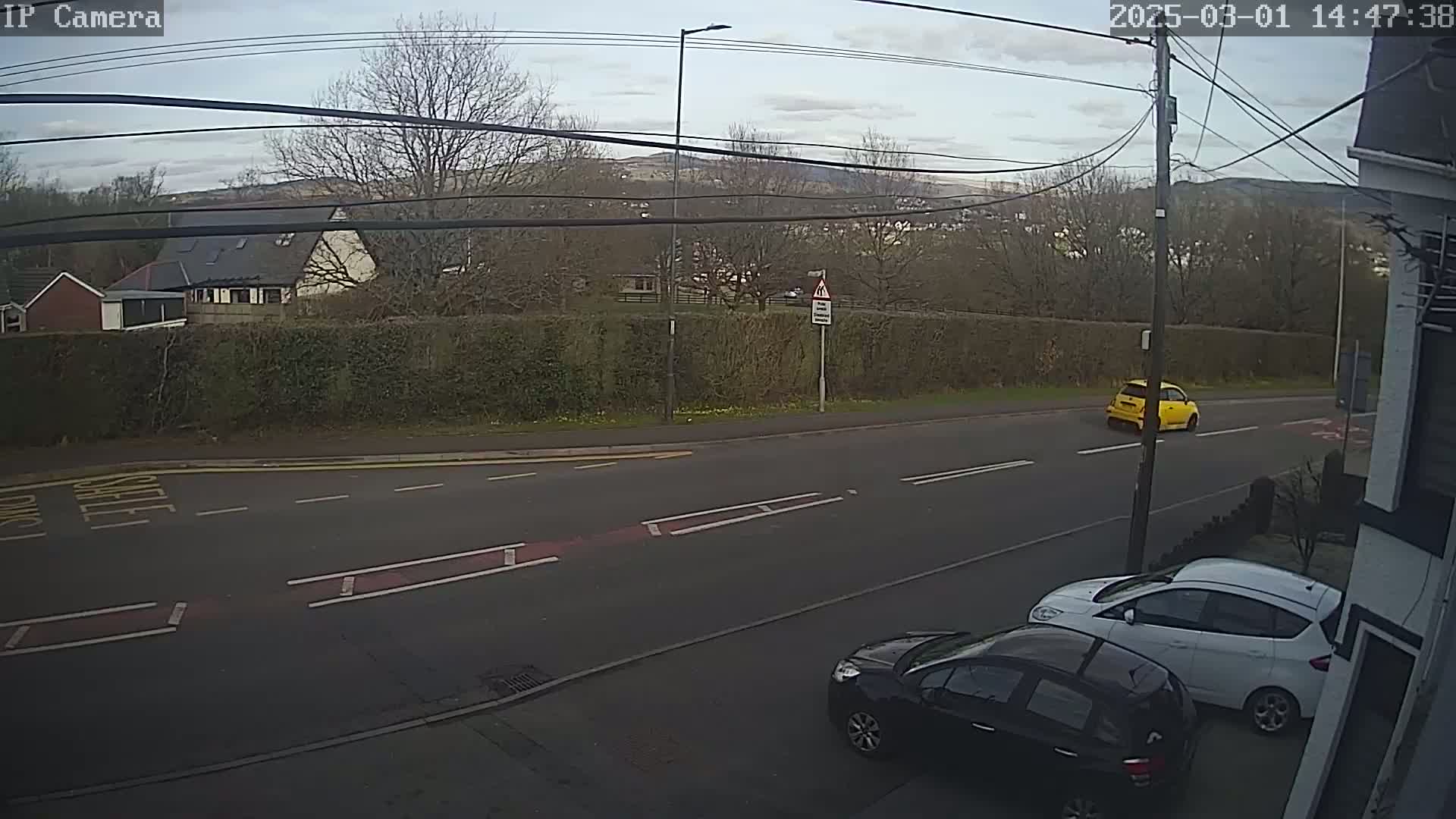 A partly cloudy day shows a road with a yellow car driving away and two parked cars, one black and one white, near a hedge and houses in the background.
