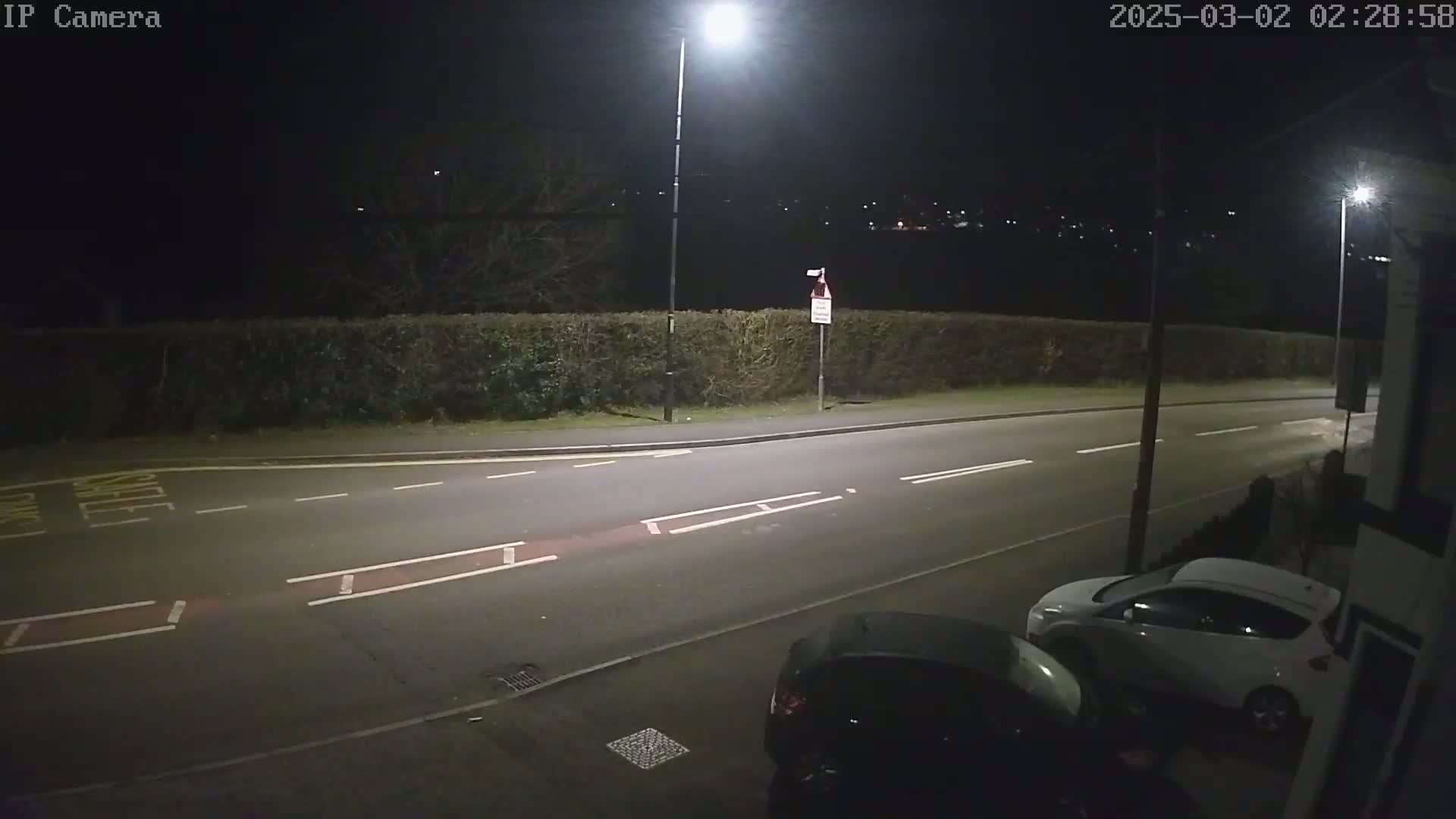 A nighttime view of a mostly empty road, bordered by a hedge, with two cars parked at the roadside under bright streetlights.