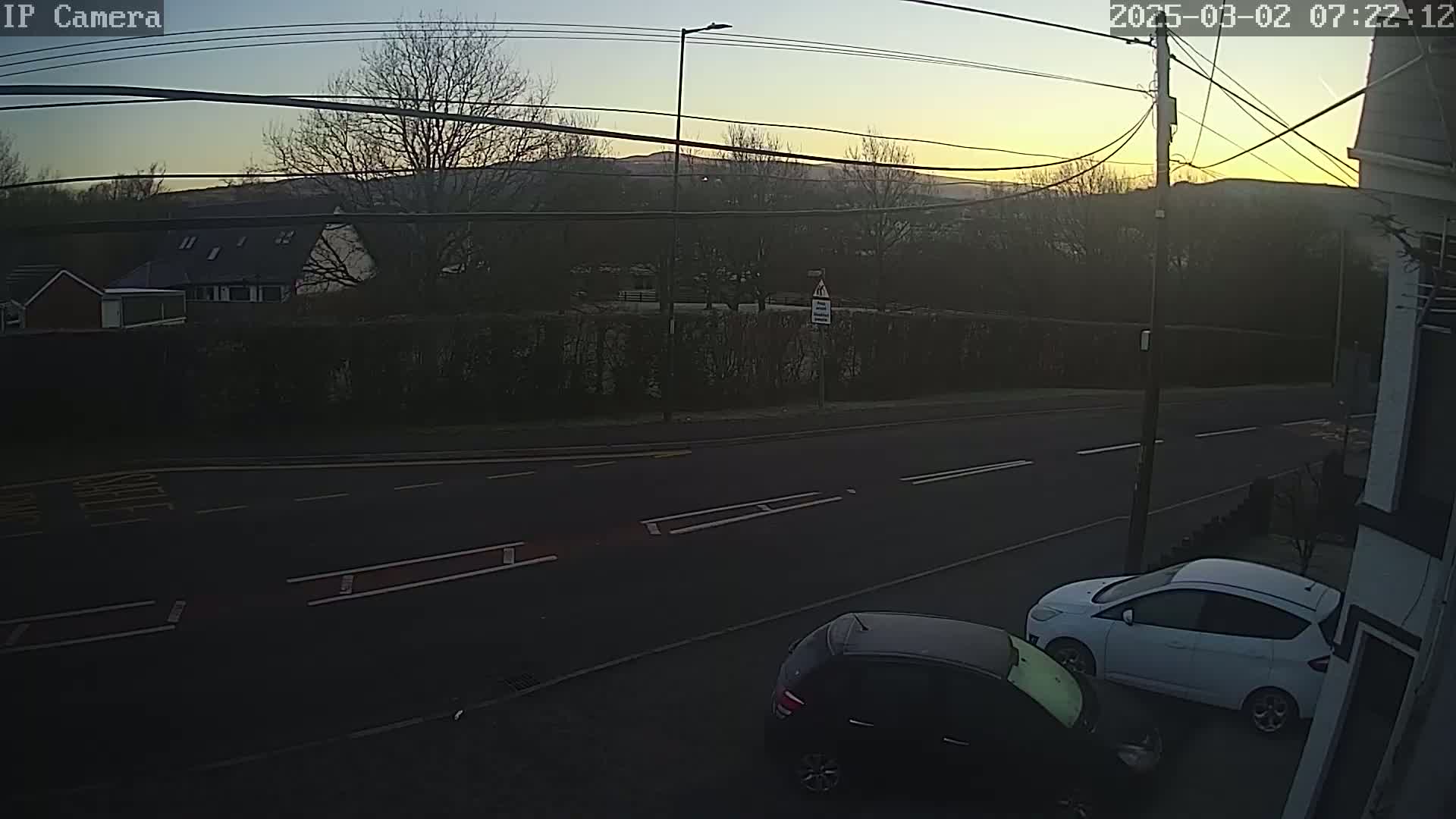 Two cars are parked on a residential street at sunrise, with a hedge and houses visible in the background.