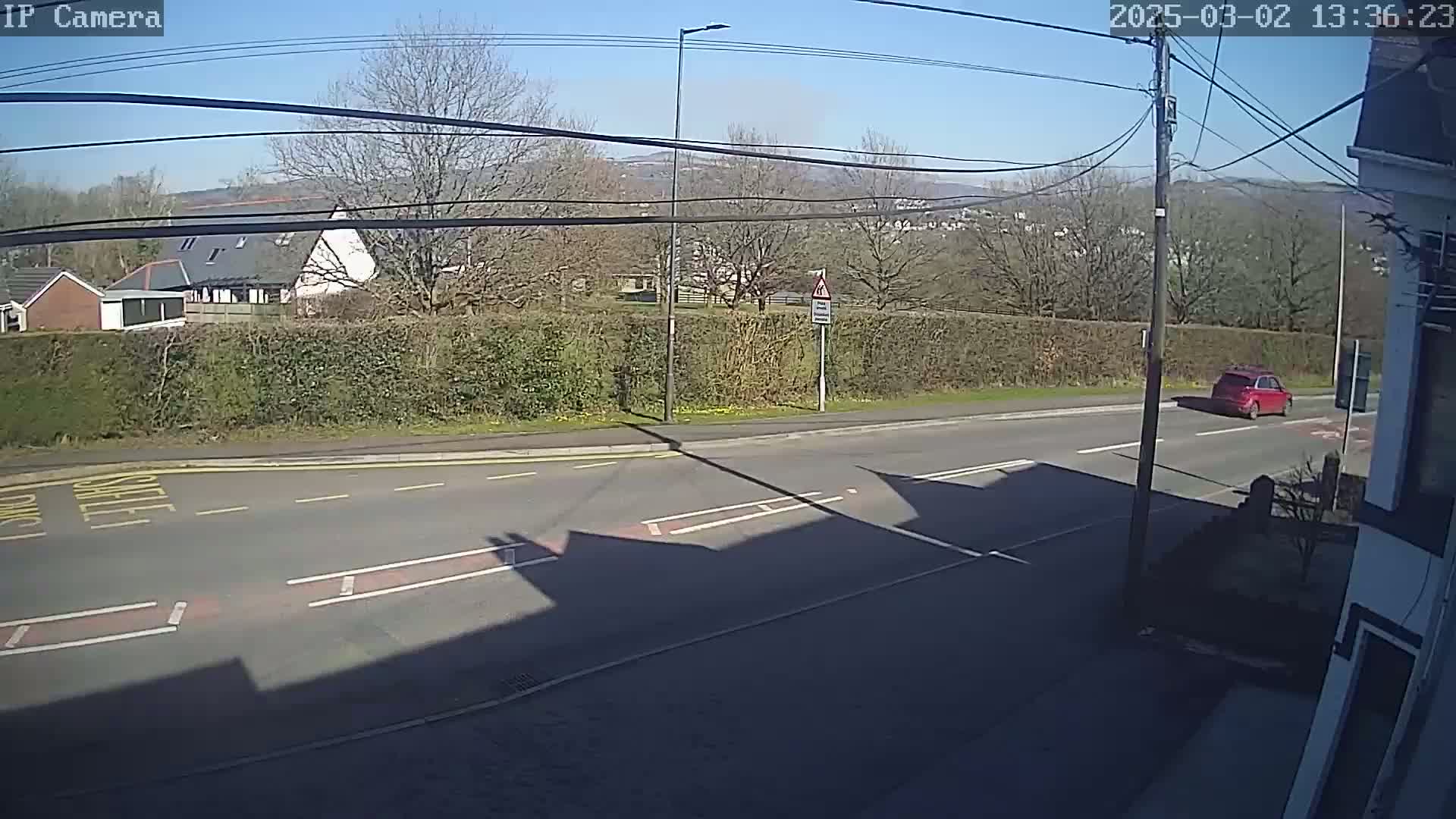 A sunny day reveals a red car driving on a road past a hedgerow, houses, and trees, with hills visible in the distance.