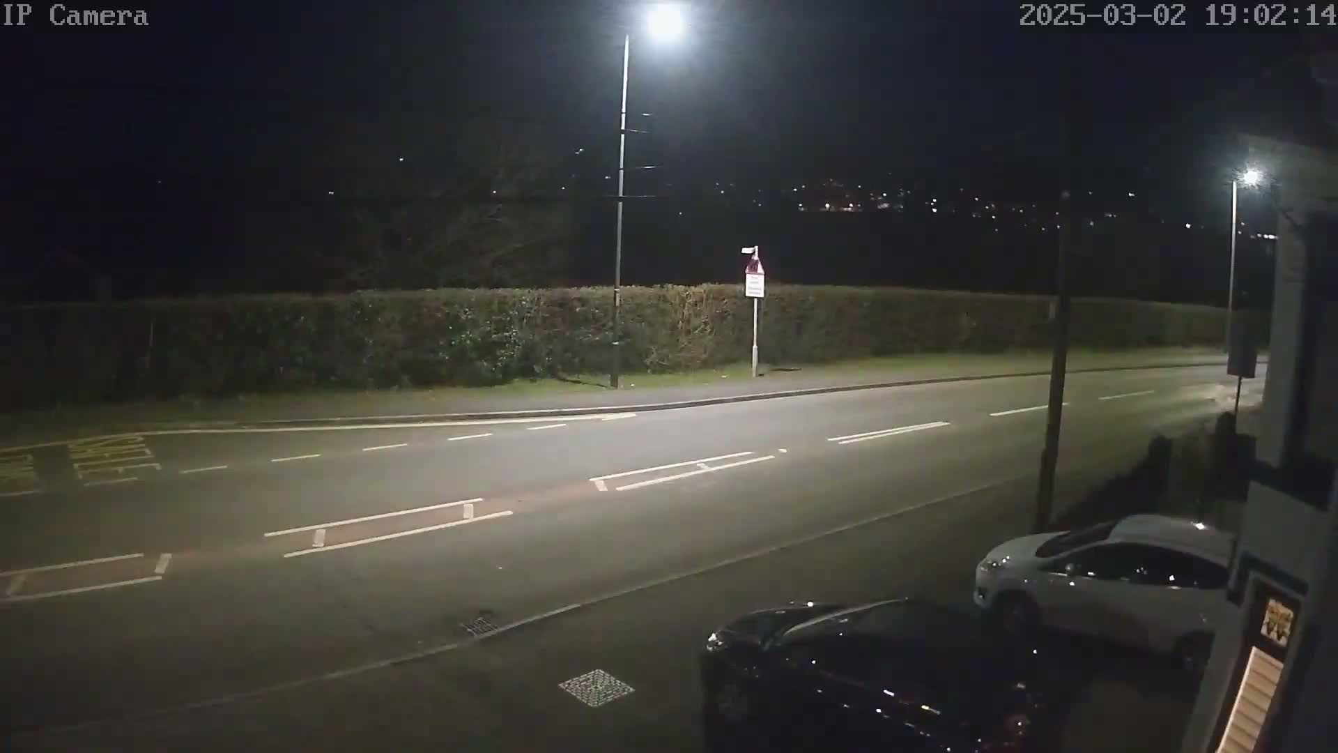 A nighttime street scene shows a road with two cars parked, lit by streetlights, with a hedge and distant lights visible in the background under a dark sky.