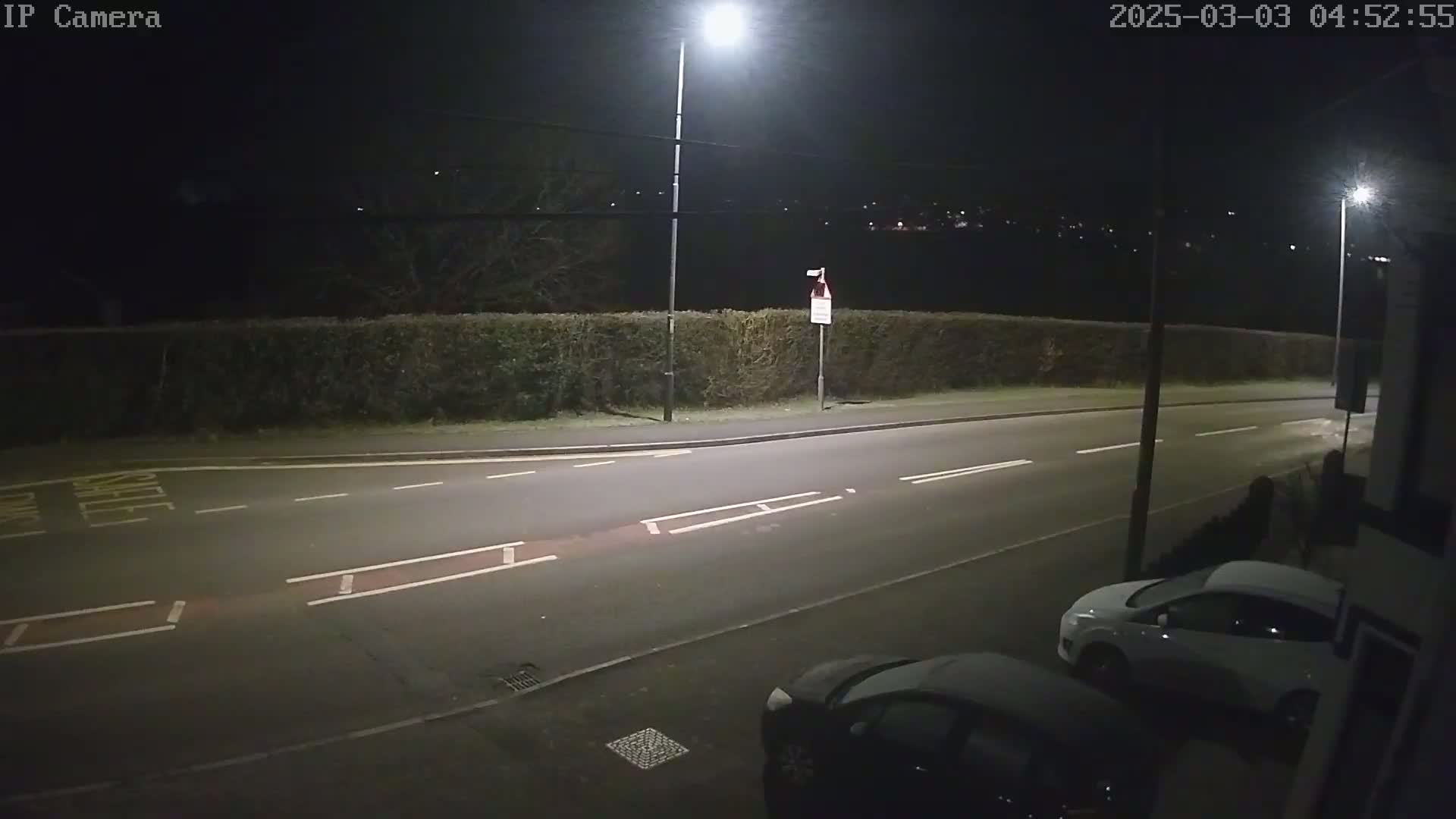 A nighttime street scene shows a road with two parked cars, a hedge, and streetlights illuminating the area.
