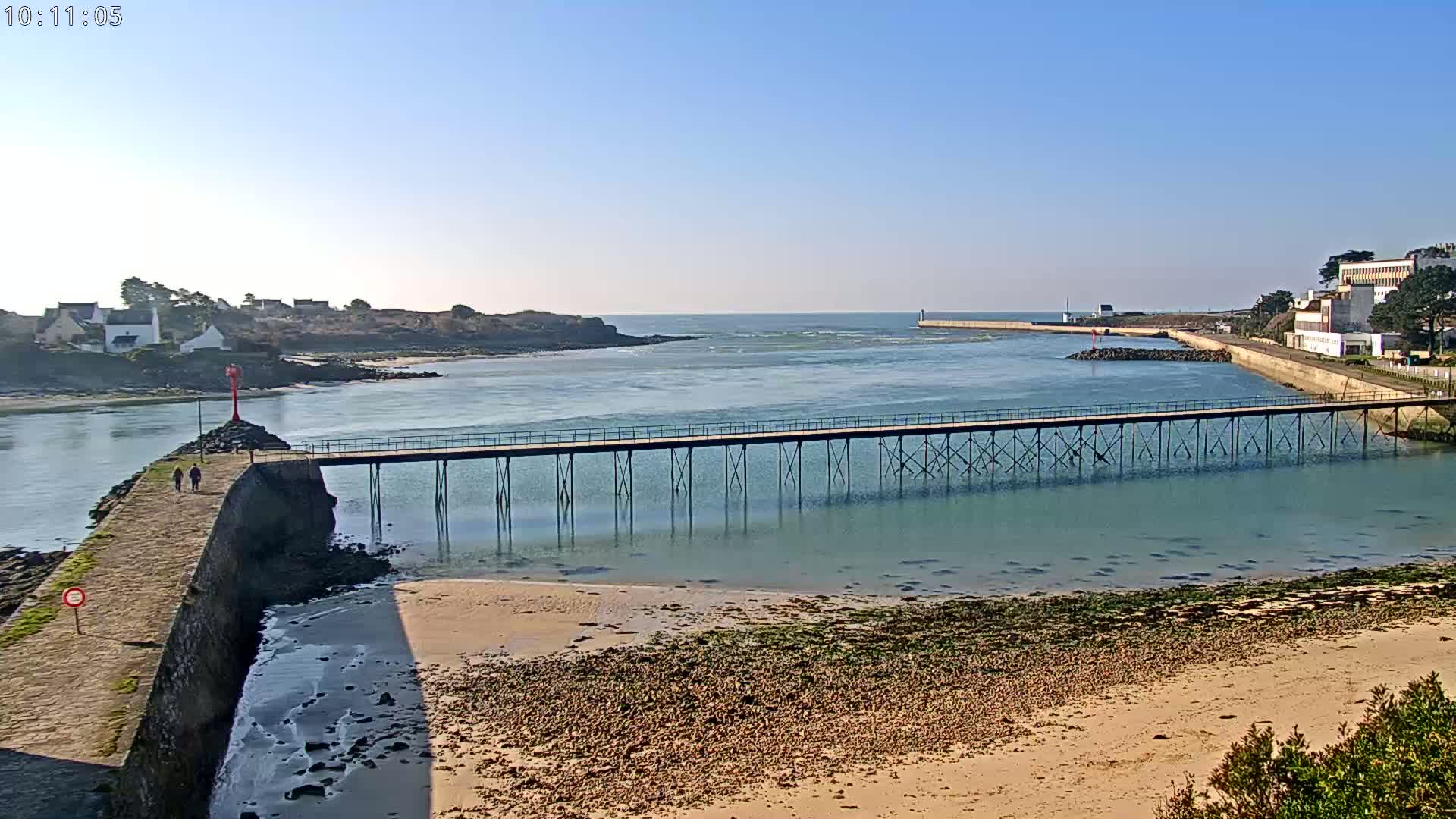 A long pier extends across calm, shallow water toward a small village and a larger building on the opposite shore under a clear, sunny sky.