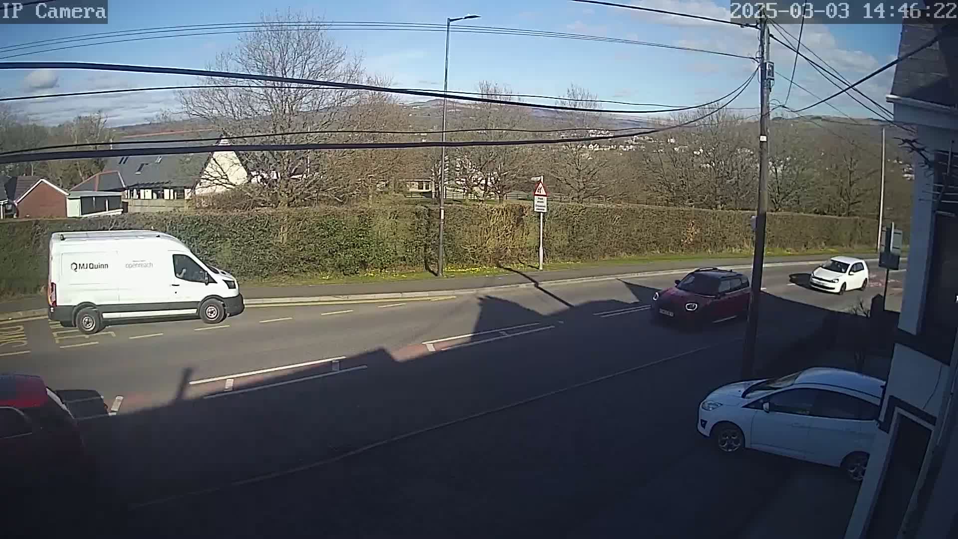 A sunny day shows a road with a white van, a red car, and a white car, all near a hedge and residential buildings, with hills visible in the background.