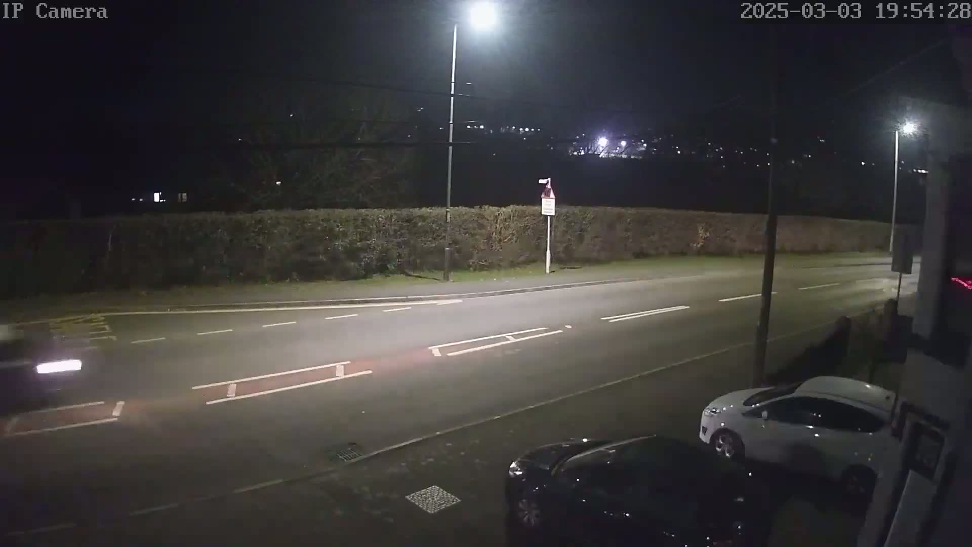 A nighttime view of a street with cars parked and driving, under bright streetlights, beside a tall hedge.