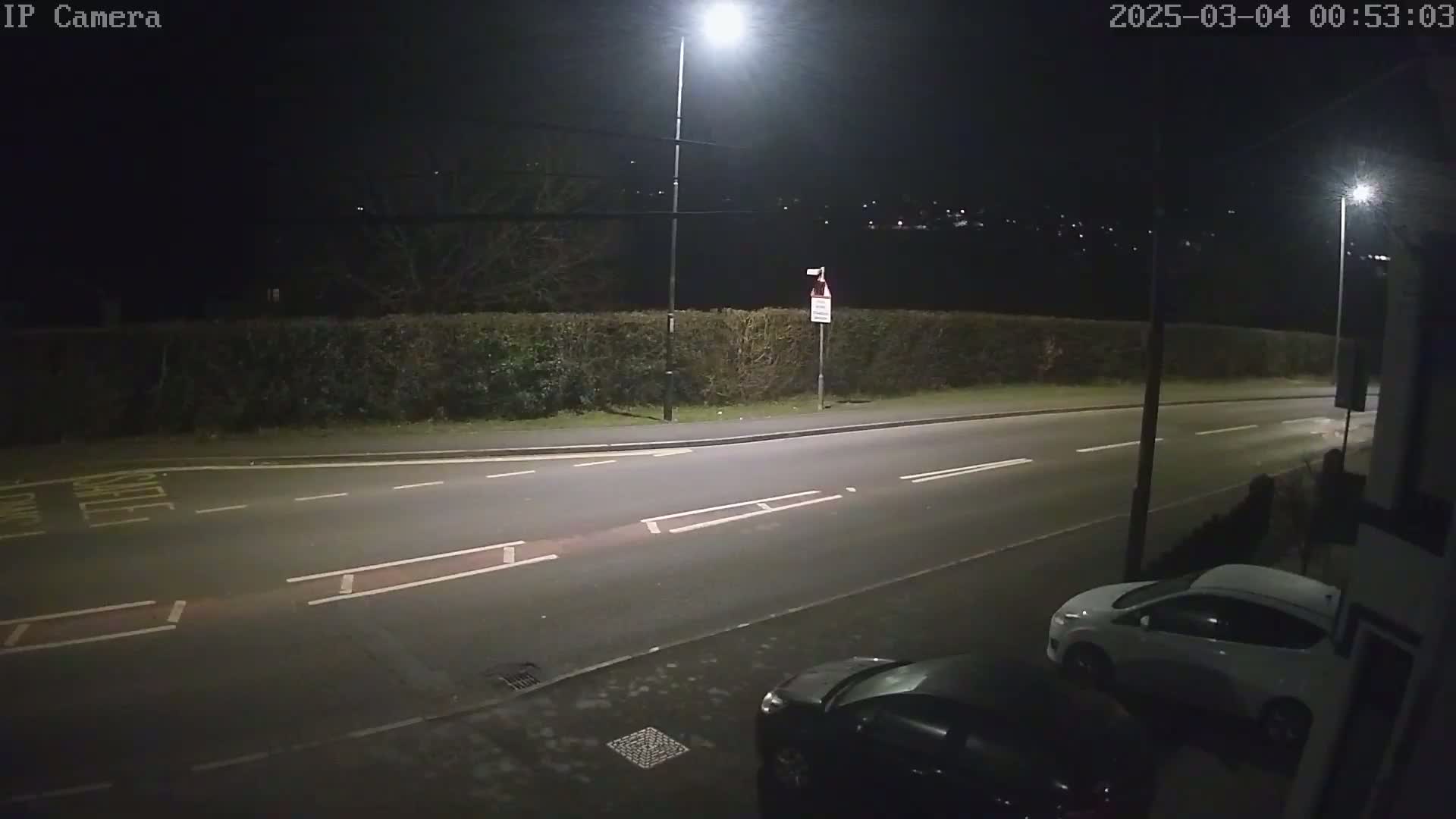 A nighttime view of a mostly empty road with cars parked on the side, illuminated by streetlights, bordered by a hedge.