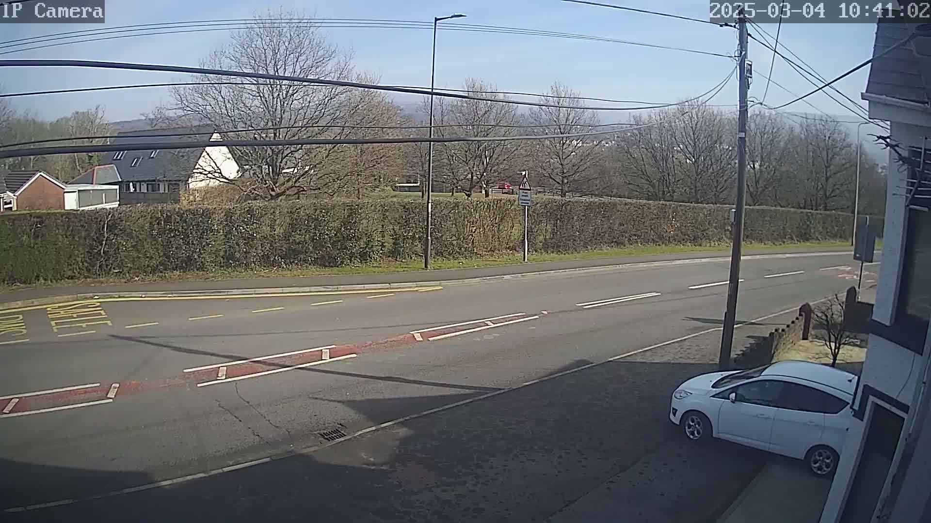 A white car is parked on the side of a road on a sunny day, with a tall hedge and buildings visible in the background.