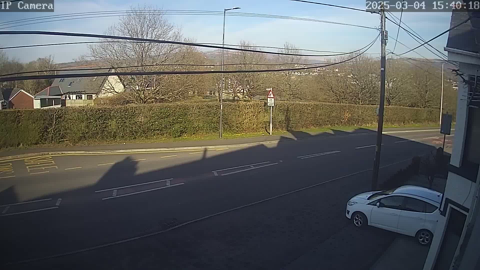 A sunny day shows a white car parked on the side of a road next to a hedge, with houses and hills visible in the background.