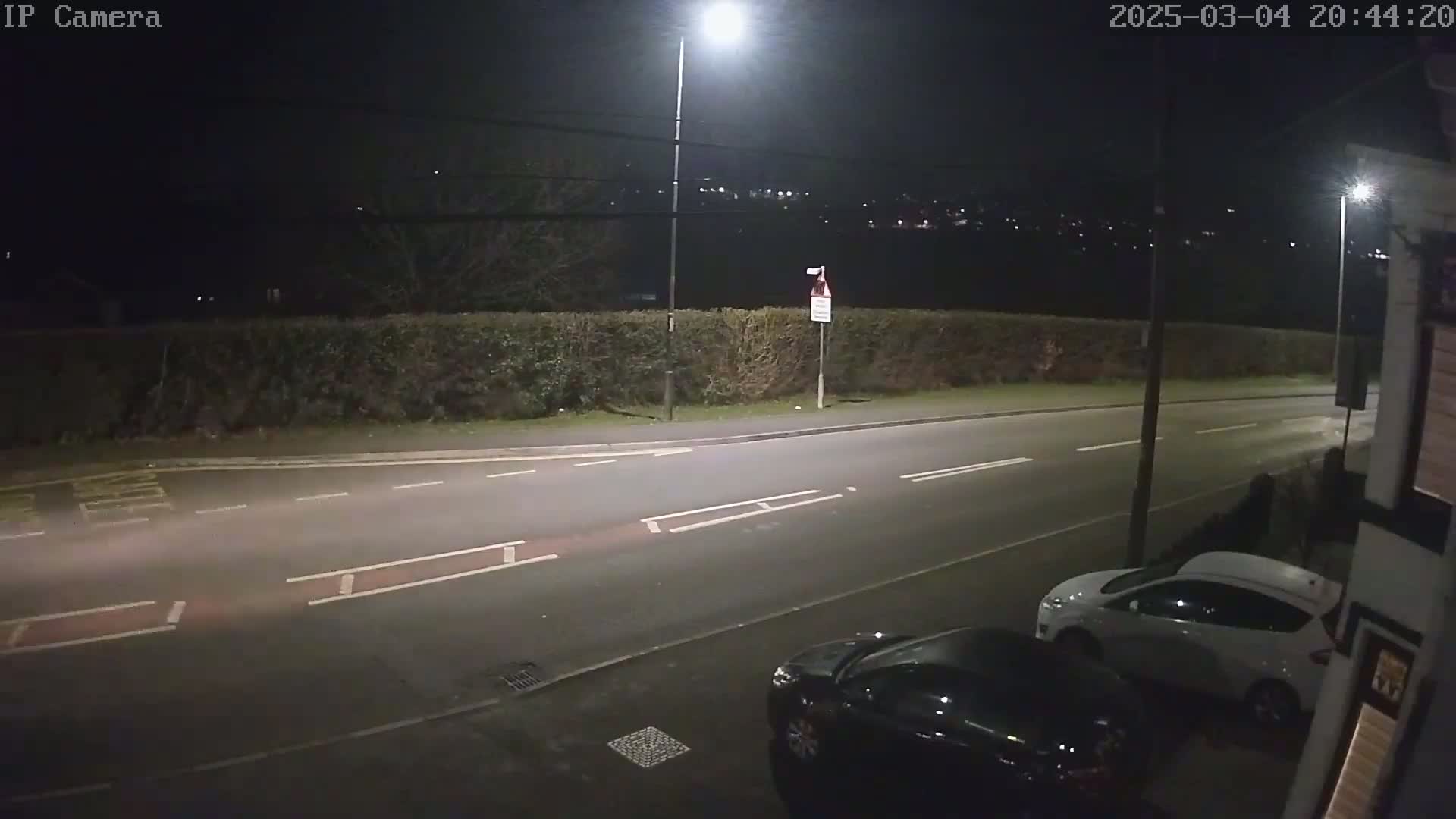 A nighttime street scene shows a road with two parked cars, a hedge, and streetlights illuminating the area under a dark sky.