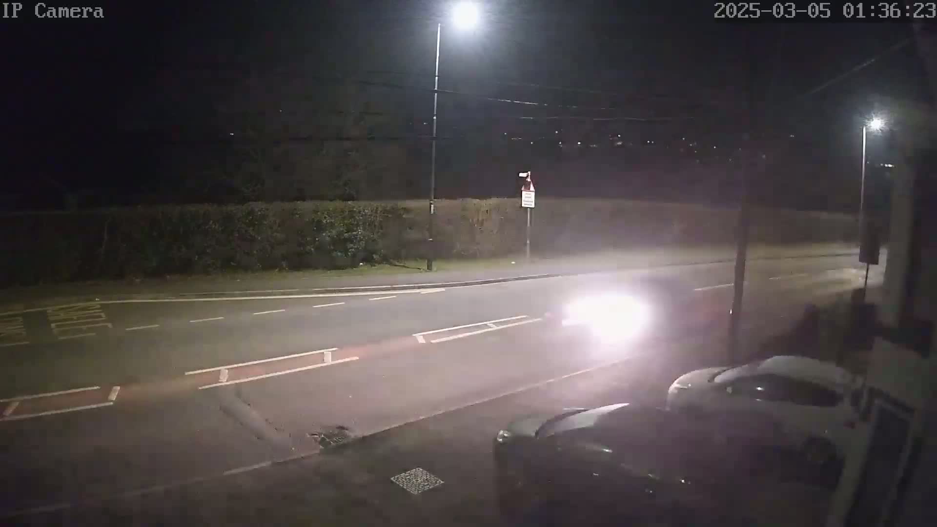 At night, a road intersection is illuminated by a streetlight, with two cars visible, one approaching and one parked nearby, under partly cloudy conditions.