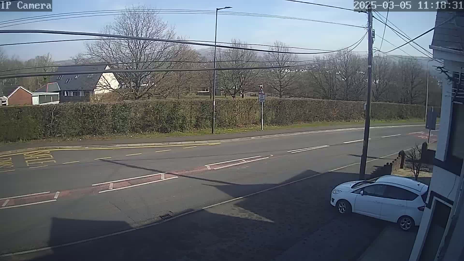 A sunny day reveals a mostly empty road with a white car parked at the side, a hedgerow across from it, and houses and trees in the distance.