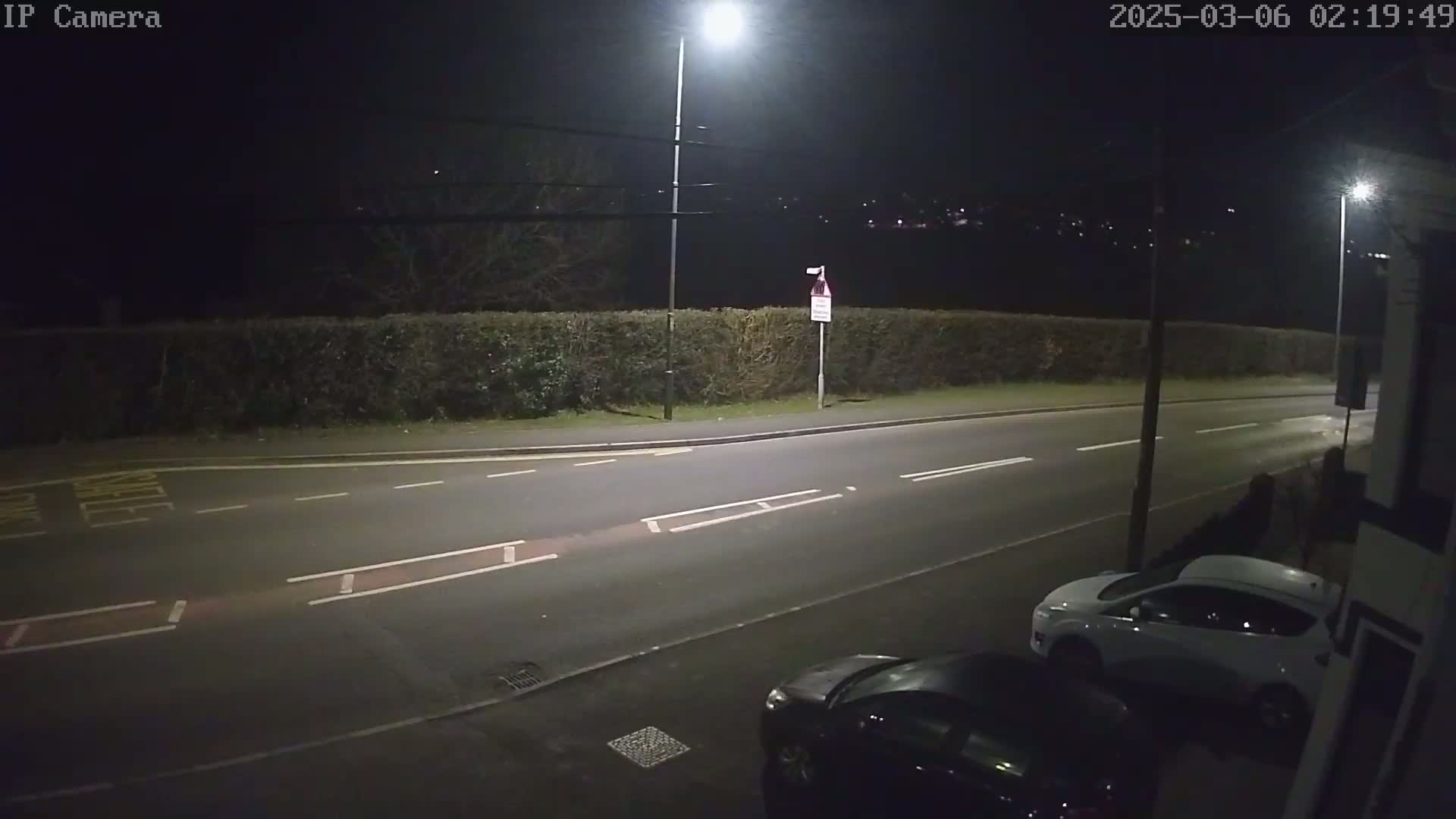 A nighttime street scene shows a road with two parked cars, a hedge, and streetlights illuminating the area.