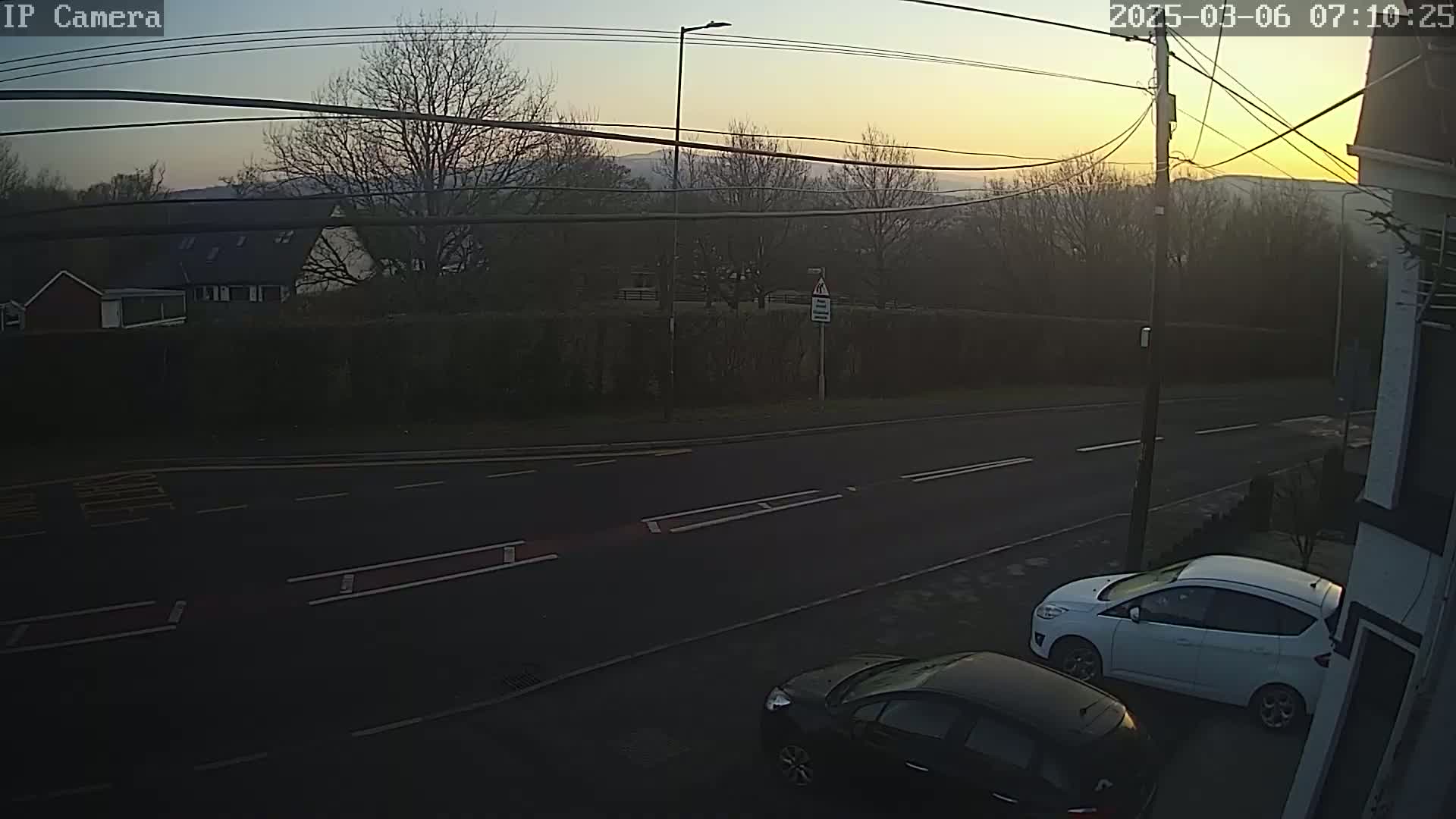 Two cars are parked on a residential street at sunrise, with a hedge and houses visible in the background.