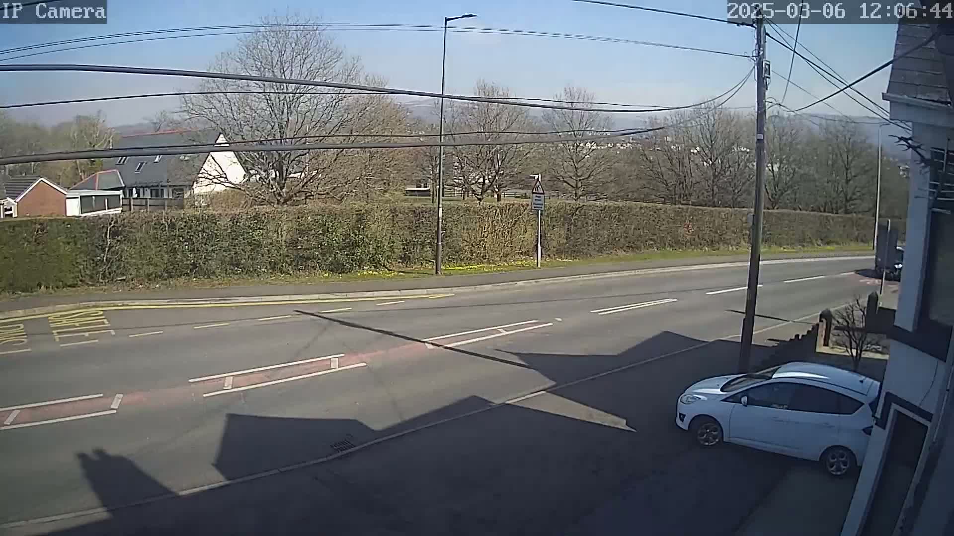A sunny day shows a view of a residential street with a white car parked near a building, a hedge, and distant hills.