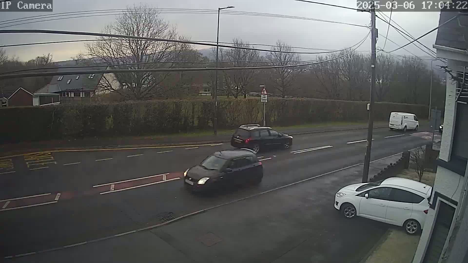 A street scene on an overcast day shows three cars and a van on a road bordered by hedges and houses, with power lines overhead.