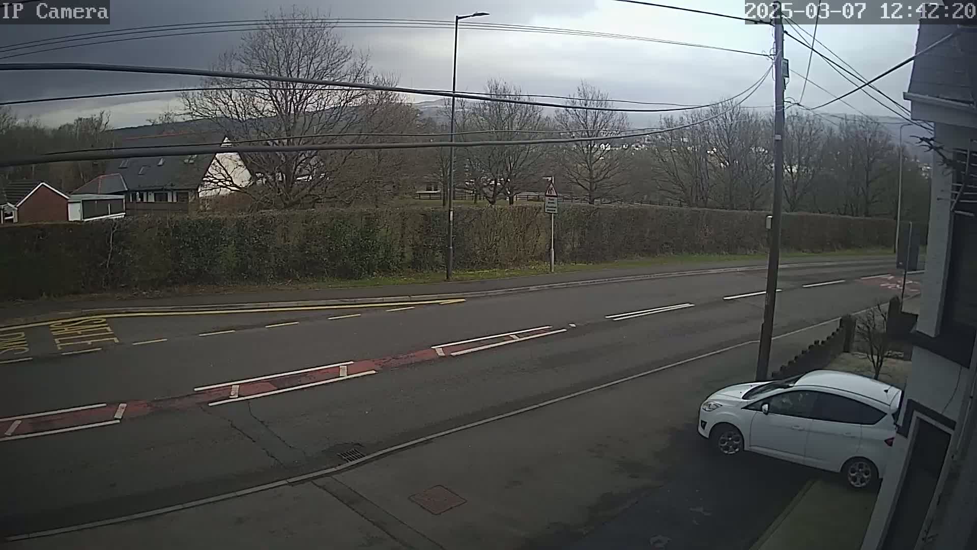 A mostly cloudy day shows a residential street with a white car parked on the side, a hedge separating the street from houses and hills in the background.