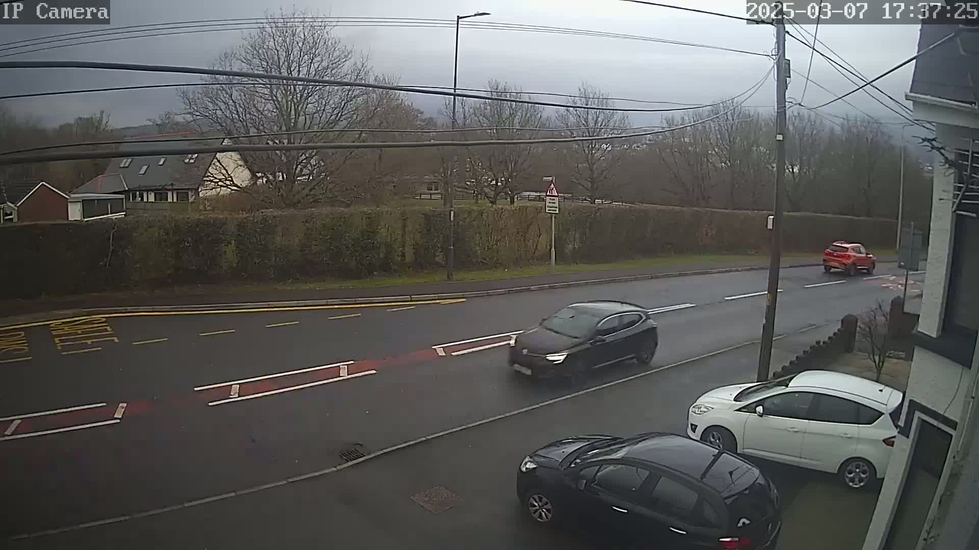A street scene on an overcast day shows three cars, two parked and one driving, on a road bordered by a hedge and houses.