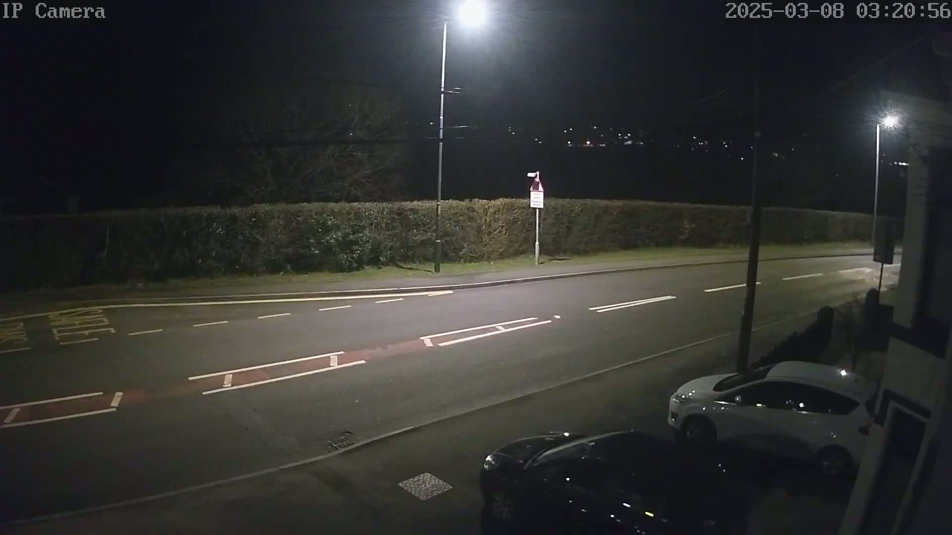 A nighttime street scene shows a road with parked cars and a hedge illuminated by streetlights.