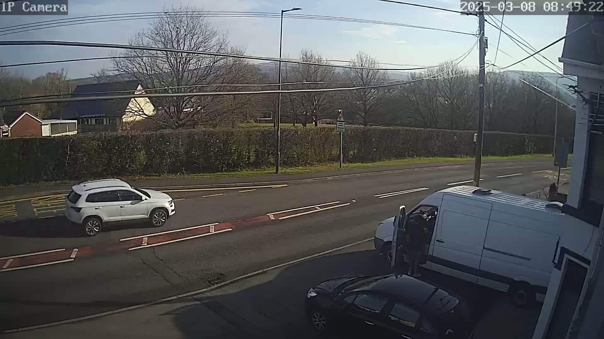 A sunny day shows a white SUV and a white van parked on a road near a hedge, with a dark-colored car partially visible in the bottom right corner.