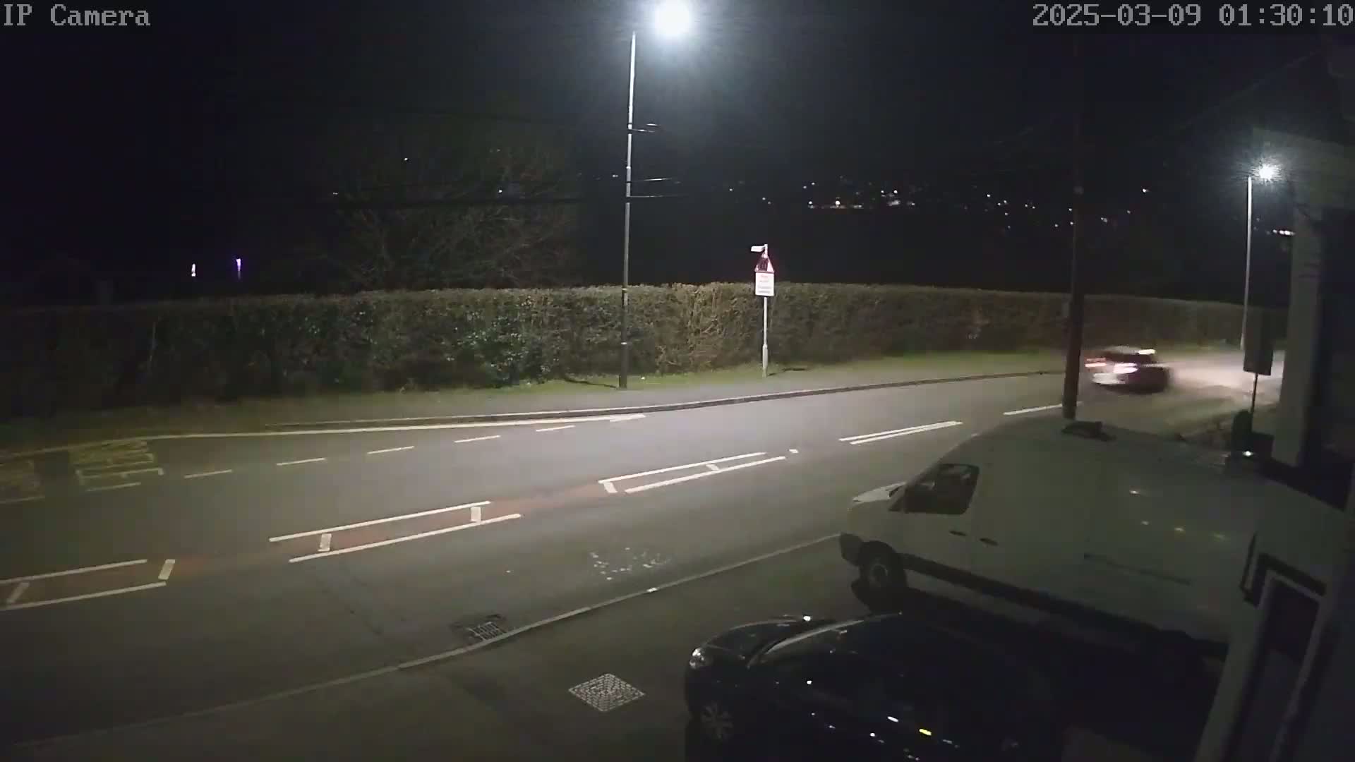 A nighttime street scene shows a white van parked near a dark-colored car, with another car driving away in the distance under streetlights.