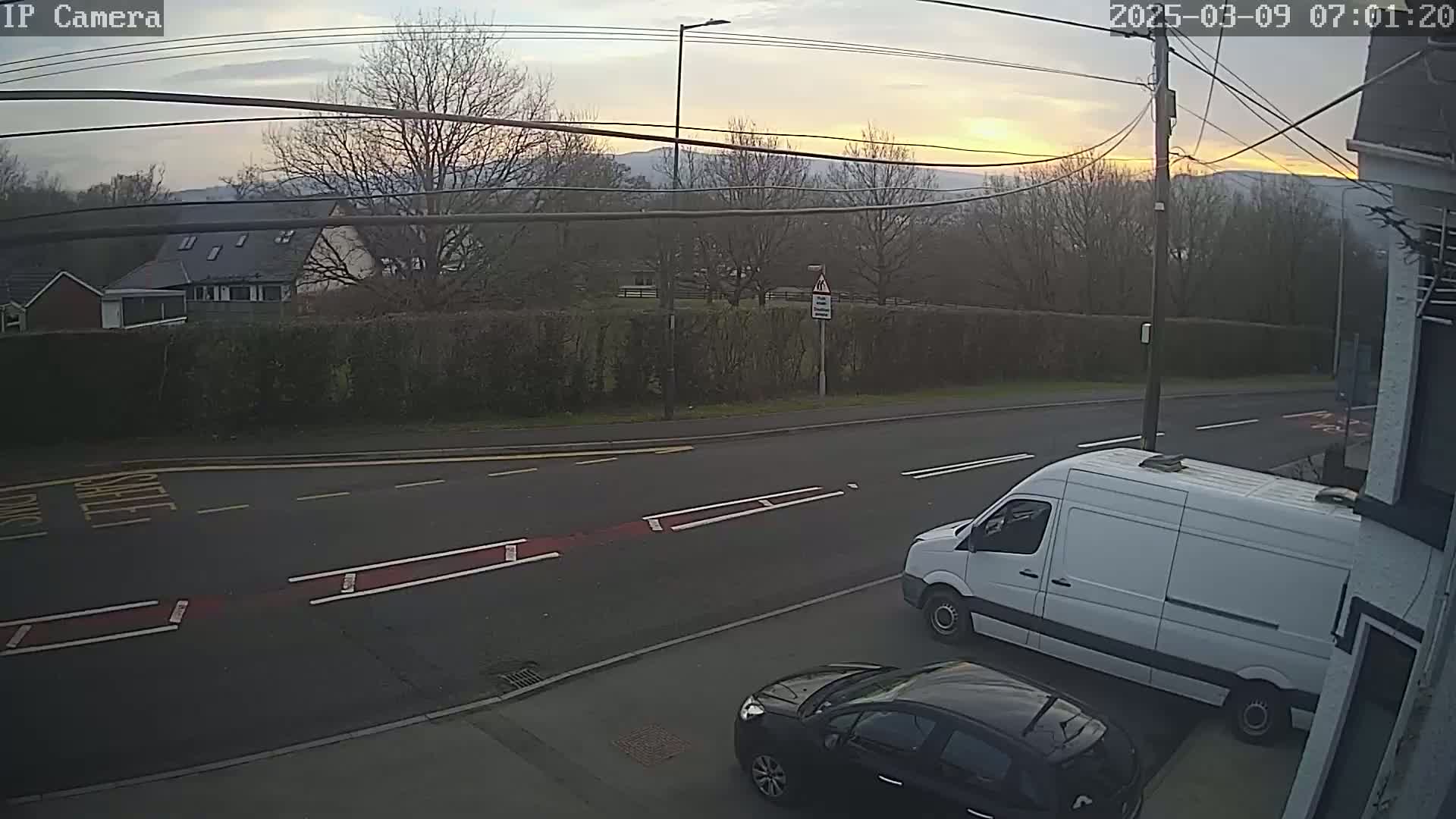A white van and a dark-colored car are parked on the side of a road at sunrise, with houses and trees visible in the background.