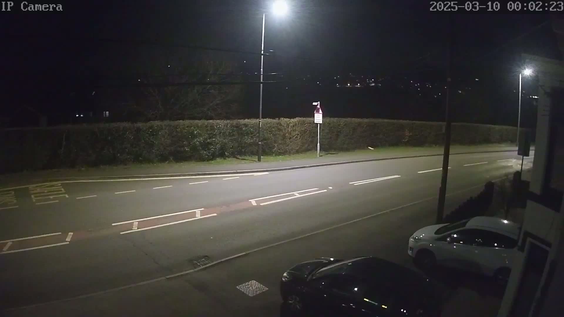 A nighttime street scene shows a road with two cars parked along the curb, illuminated by streetlights, with a hedge and distant houses visible under a dark sky.