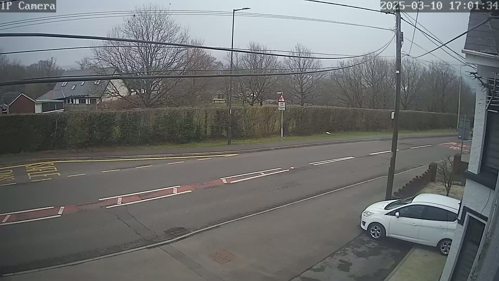 A mostly overcast day shows a residential street with a white car parked on the side, a hedge across the street, and some buildings in the background.