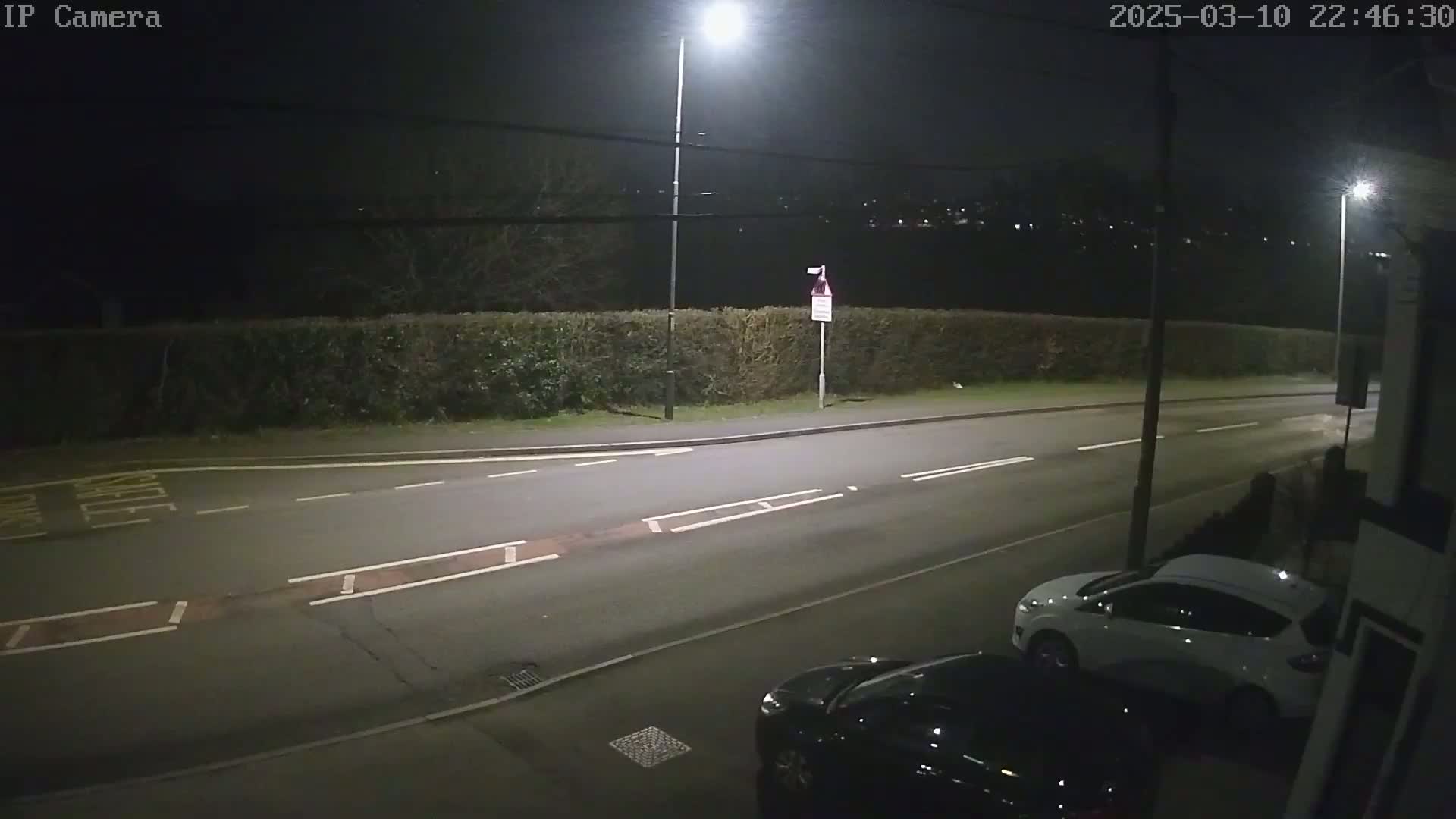 A nighttime street scene shows a road with two cars parked, a hedge, and streetlights illuminating the area under a dark sky.