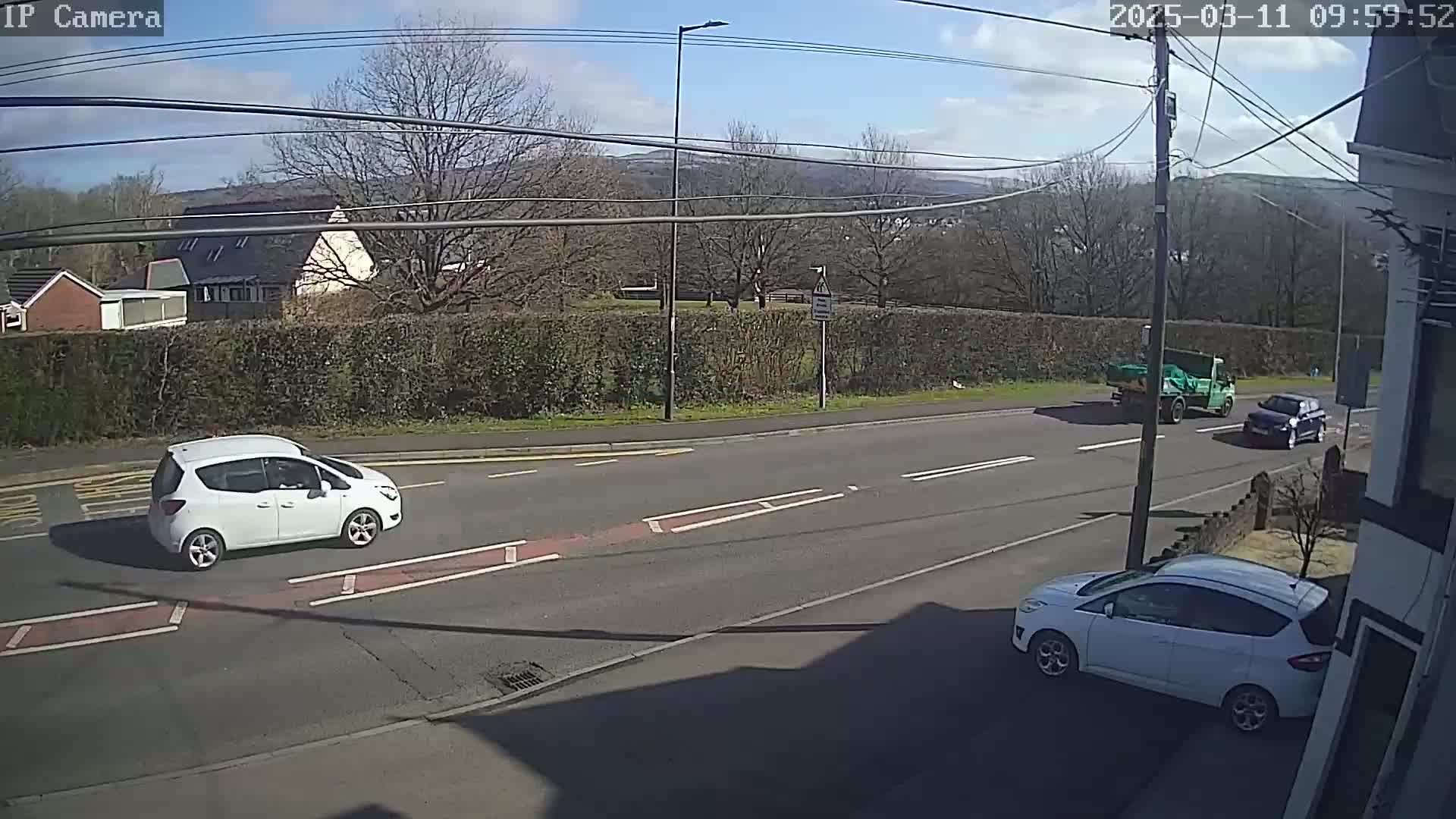 A sunny day shows a road with three cars, one of which is a white hatchback, and a green truck; all are moving on the road beside a hedge and houses.