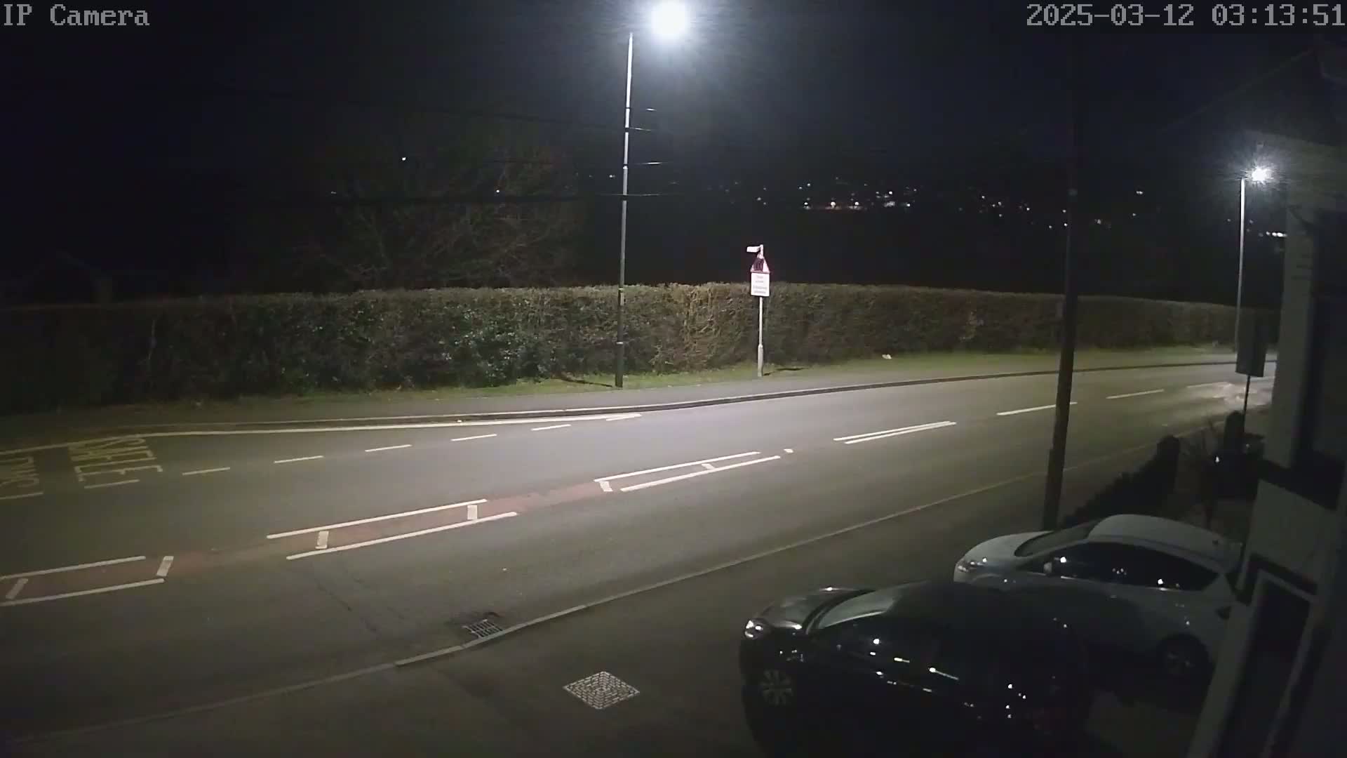 A nighttime street scene shows two parked cars and a hedge along a road lit by streetlights.