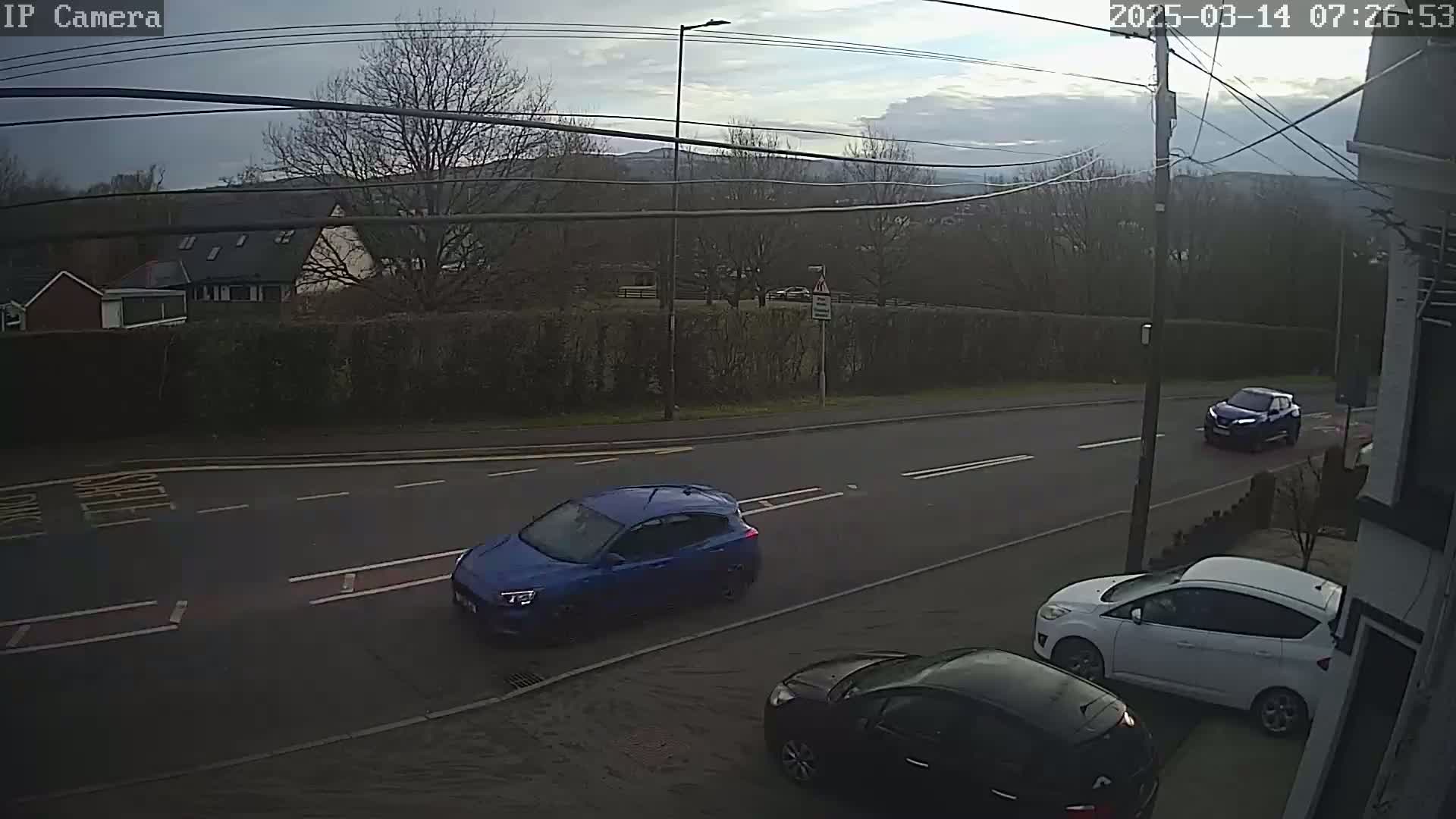 Three cars are parked and driving on a residential street under an overcast sky.