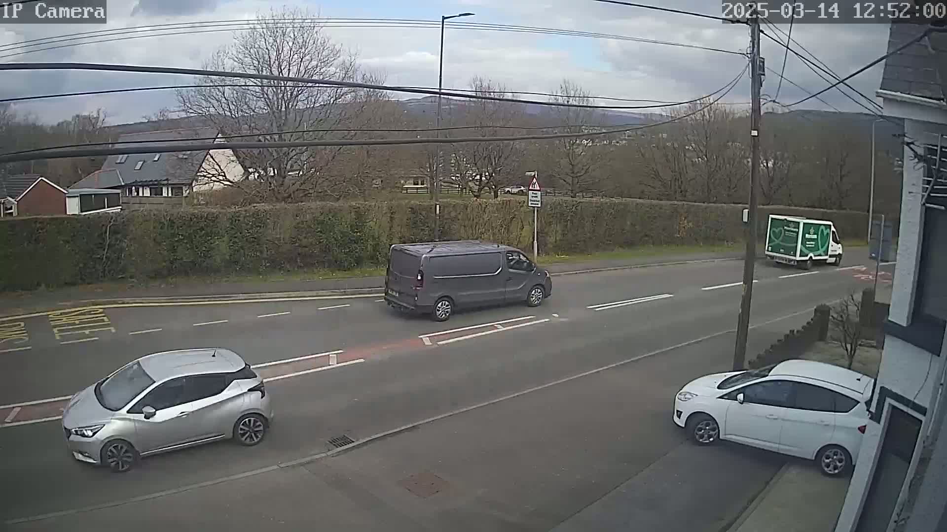 A gray van, a silver car, and a white car are parked or driving on a residential street under a partly cloudy sky.