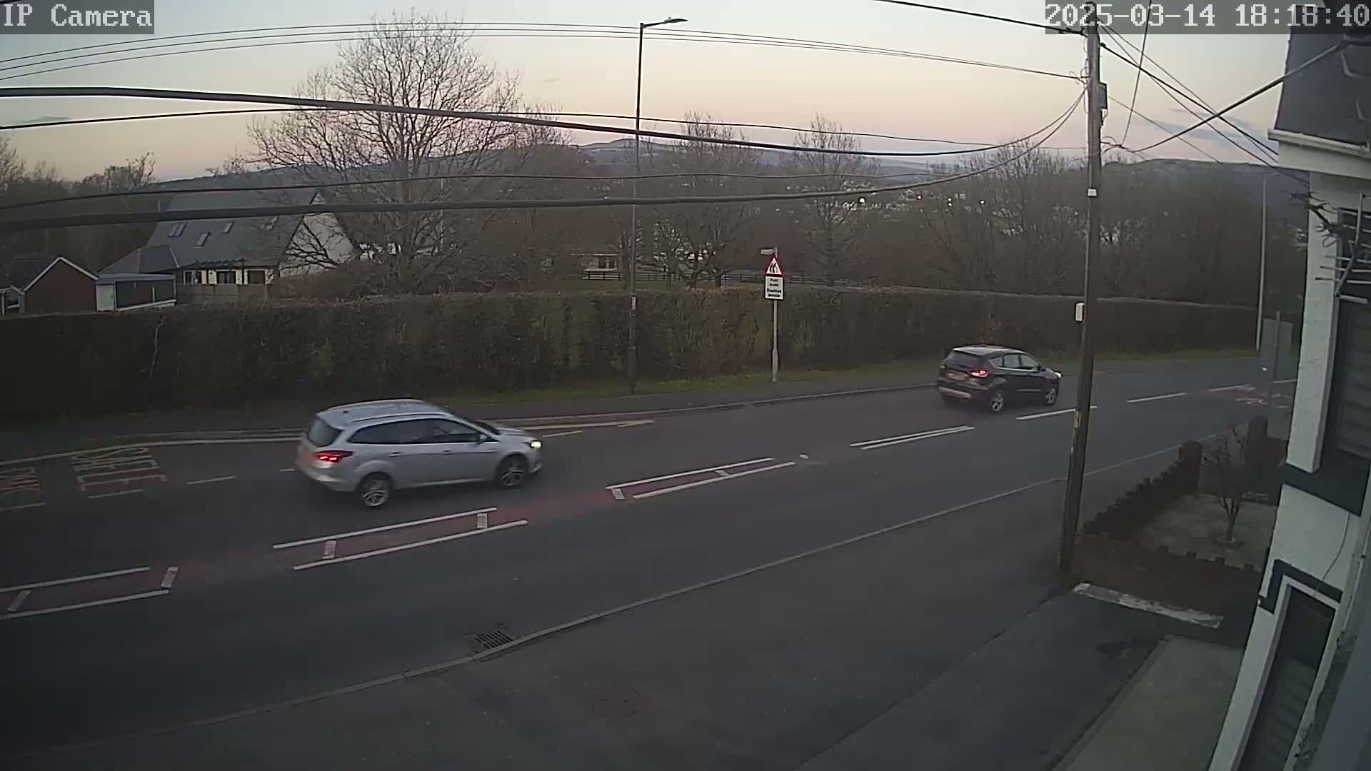 Two cars are driving on a road at dusk, past a hedgerow and houses, with hills visible in the background.